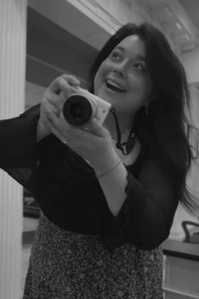 A woman taking a selfie with a digital camera, smiling and looking happy, in a room with a mirror and some furniture.