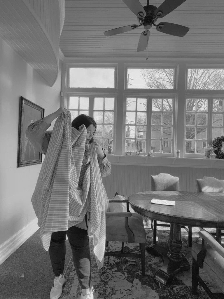 A woman with long hair, wearing a jacket, is standing in a dining room, holding a large striped cloth or blanket. The room has large windows letting in natural light, a round wooden table, and several chairs. There's a ceiling fan and framed artwork on the wall.
