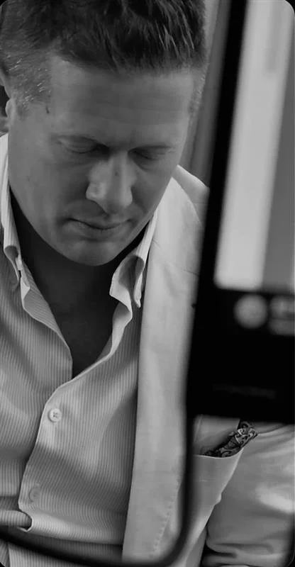 A man in a light-colored blazer and collared shirt looking downward, partially obscured by a vertical object in the foreground.