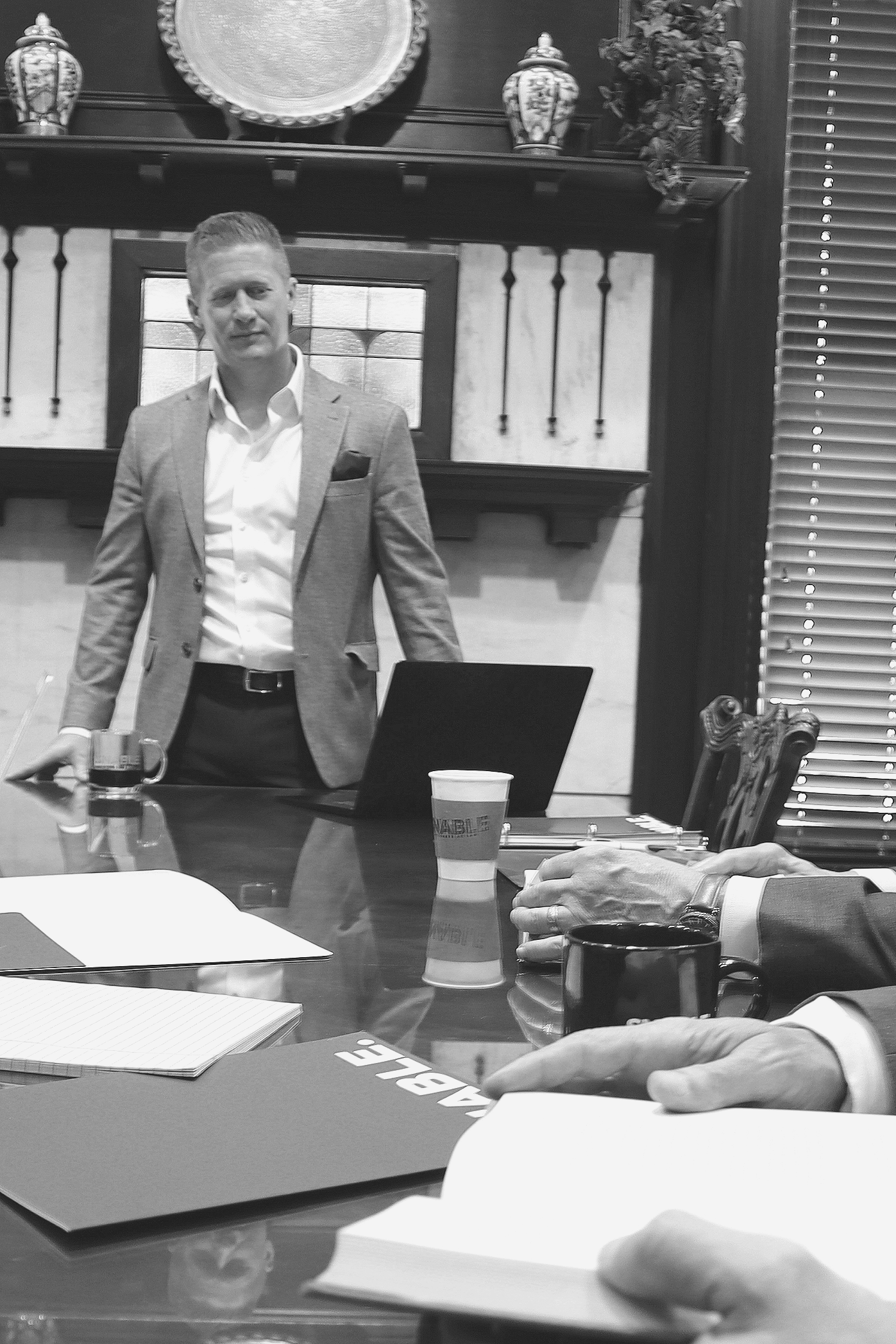 A man in a suit standing in a conference room with a table, glasses, notebooks, and hands of other people visible on the table, in black and white.