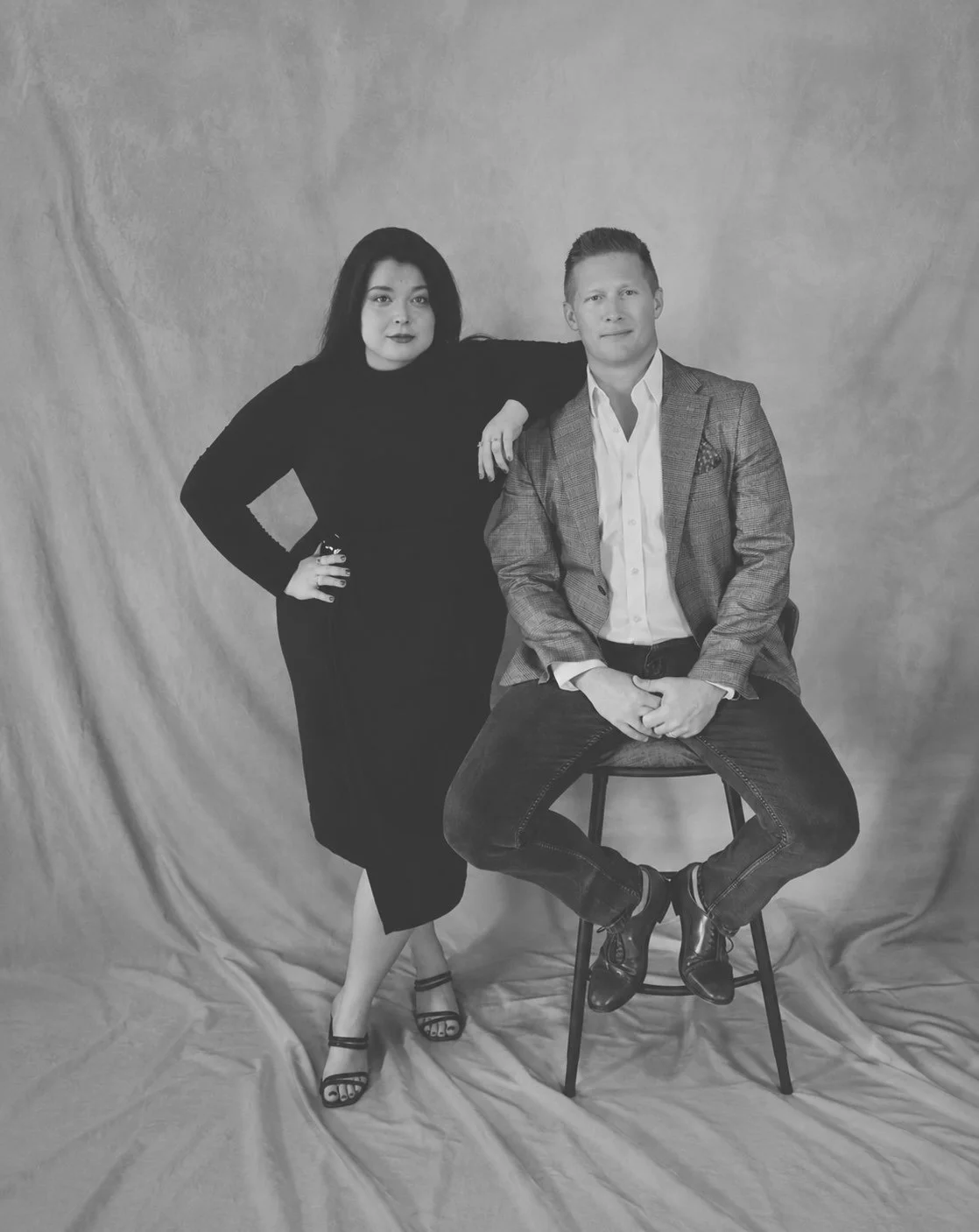A black-and-white photo of a woman and man posing together against a plain backdrop. The woman stands with her hand on her hip, wearing a long dark dress and high heels. The man sits on a stool with his hands clasped, dressed in a blazer, dress shirt, and jeans.