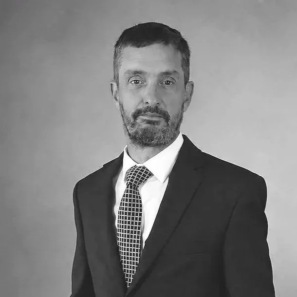 A black-and-white photo of a man in a suit and tie, with short hair and a beard, looking at the camera with a serious expression.