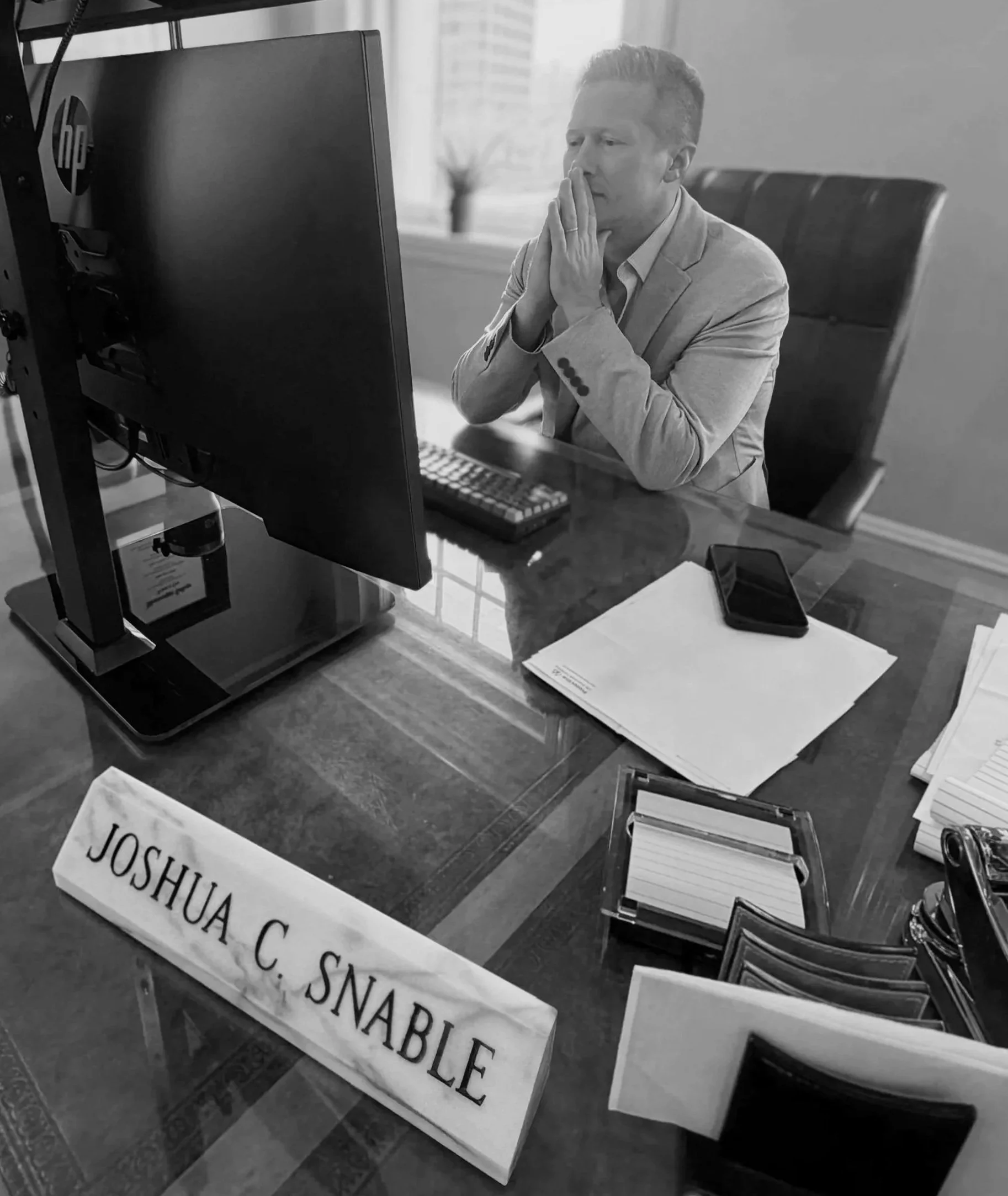 Black and white photo of a man in a suit seated at a desk, looking at a computer screen with a pensive expression, hands clasped near his face, surrounded by papers and signs.