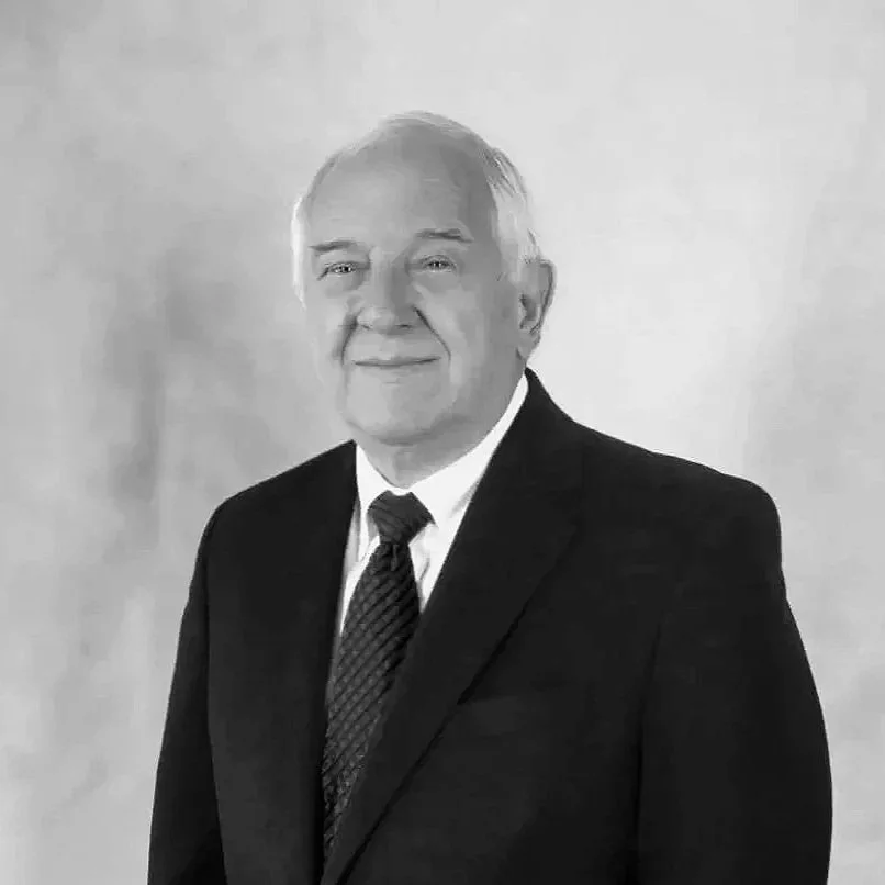 A black and white portrait of an elderly man wearing a suit and tie, smiling slightly, with a plain background.