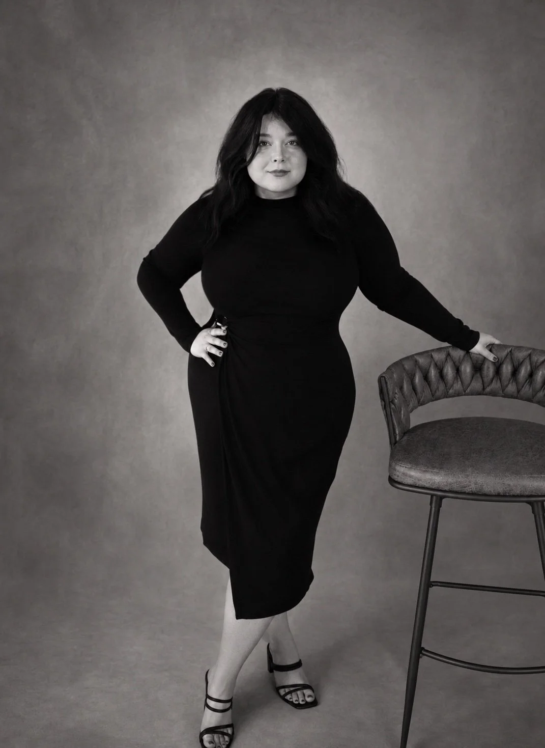 A woman standing next to a tall bar stool with her hand resting on the back of the chair. She has dark, wavy hair and is wearing a black dress and black high-heeled sandals. The background is plain and studio-style.