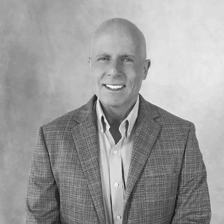 A black-and-white photo of a smiling man with short hair, wearing a checkered blazer over a collared shirt, standing against a plain background.