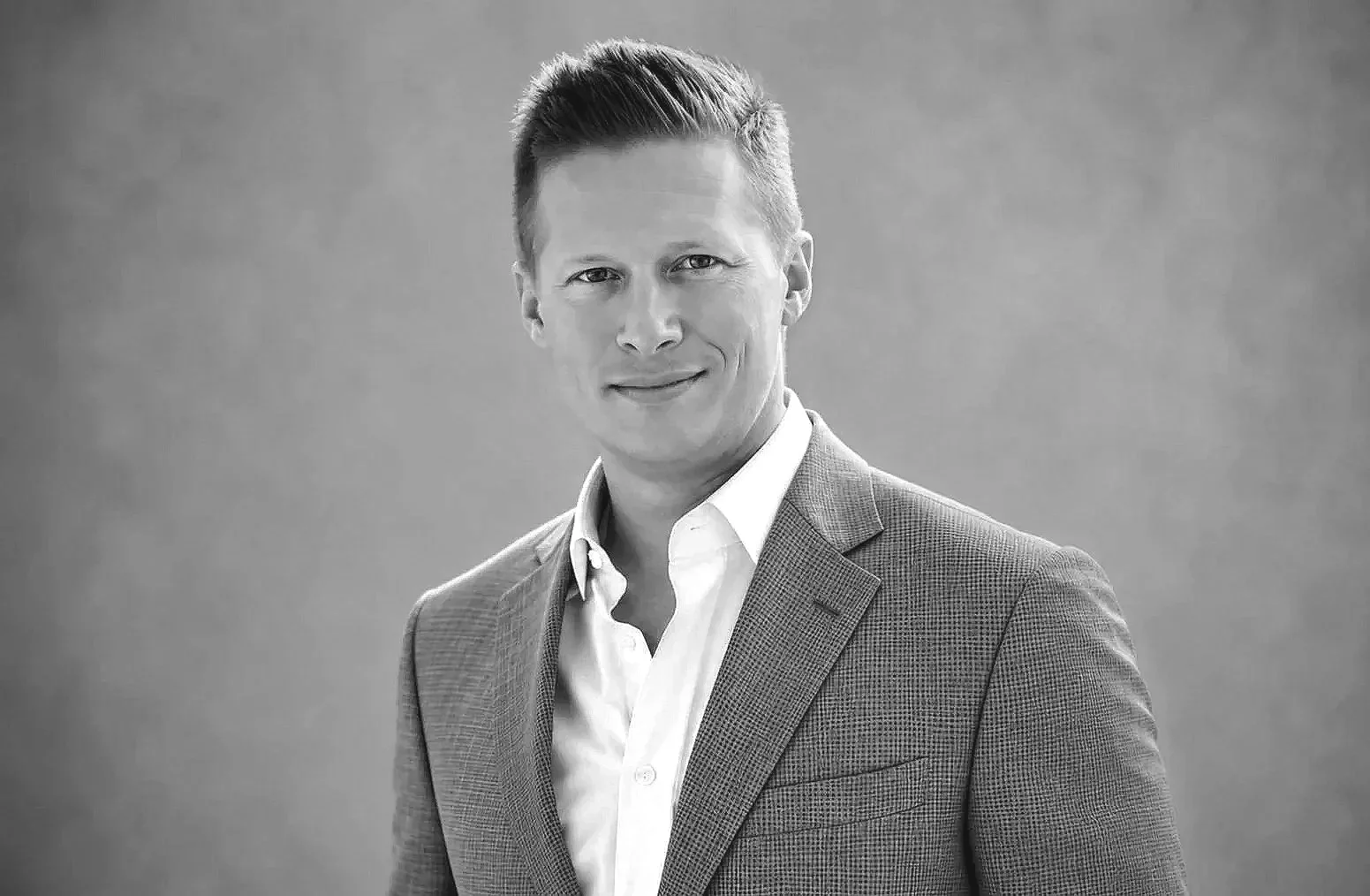 Black and white professional portrait of a man in a suit, smiling, with short, styled hair, standing against a plain background.