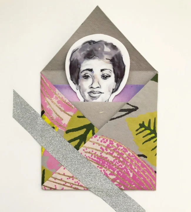 Sticker with Audre Lorde’s portrait peaks out of a floral patterned origami envelope adhered to a white board with silver sparkle washi tape.