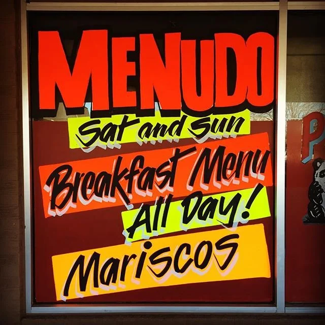 Colorful sign advertising a menu for Saturday and Sunday with breakfast and mariscos, featuring red, yellow, and black text.