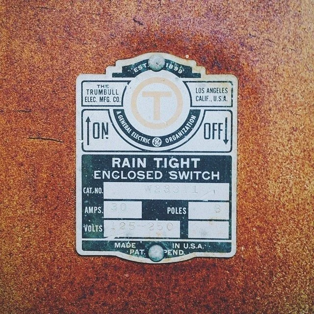 Metal identification plate for a rain-tight enclosed switch, mounted on a rusted surface, issued by Trumbull Electric Manufacturing Company in Los Angeles, California, U.S.A.