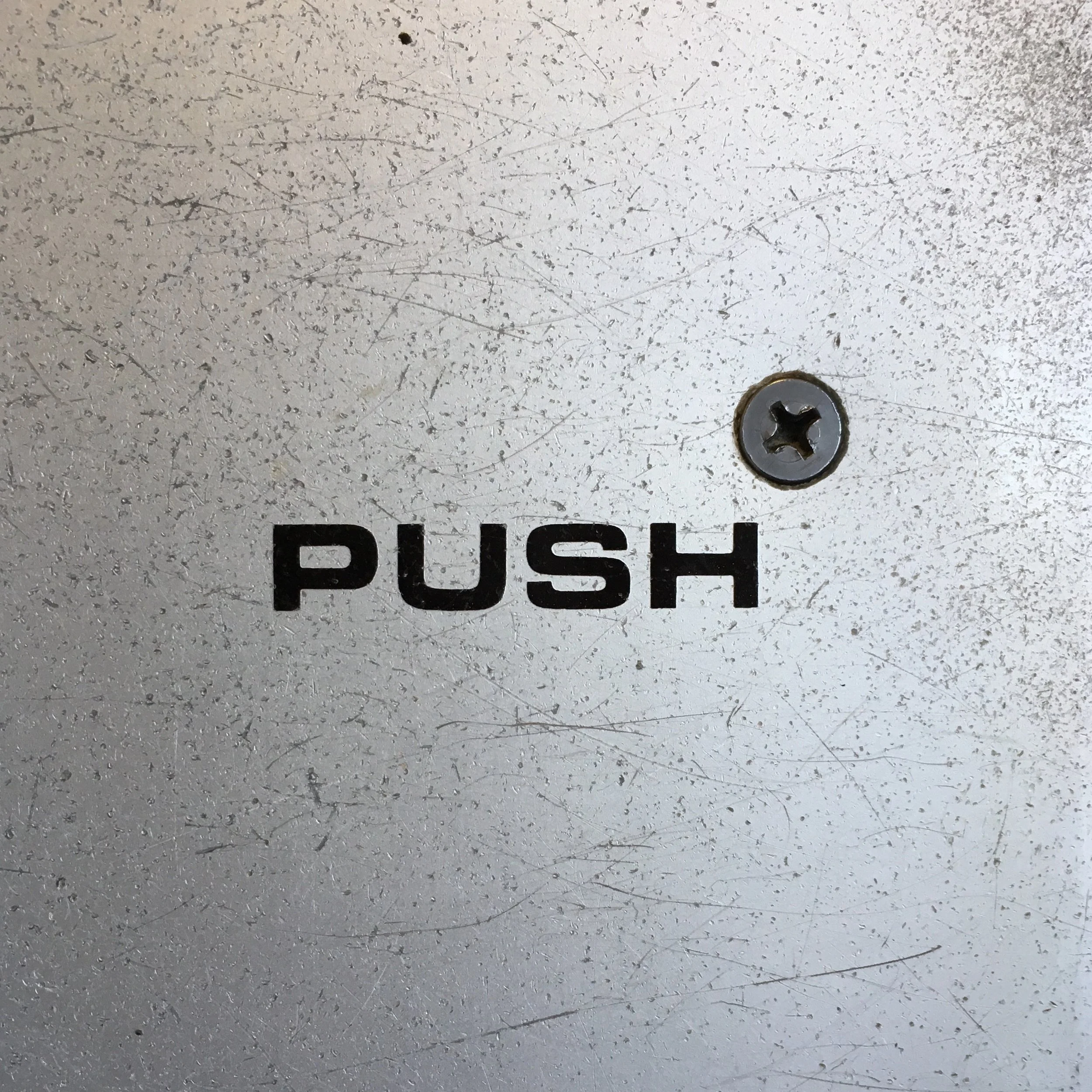 Close-up of a textured white surface with scratches, small holes, a partially visible black label with the word 'PUSH', and a metal screw.