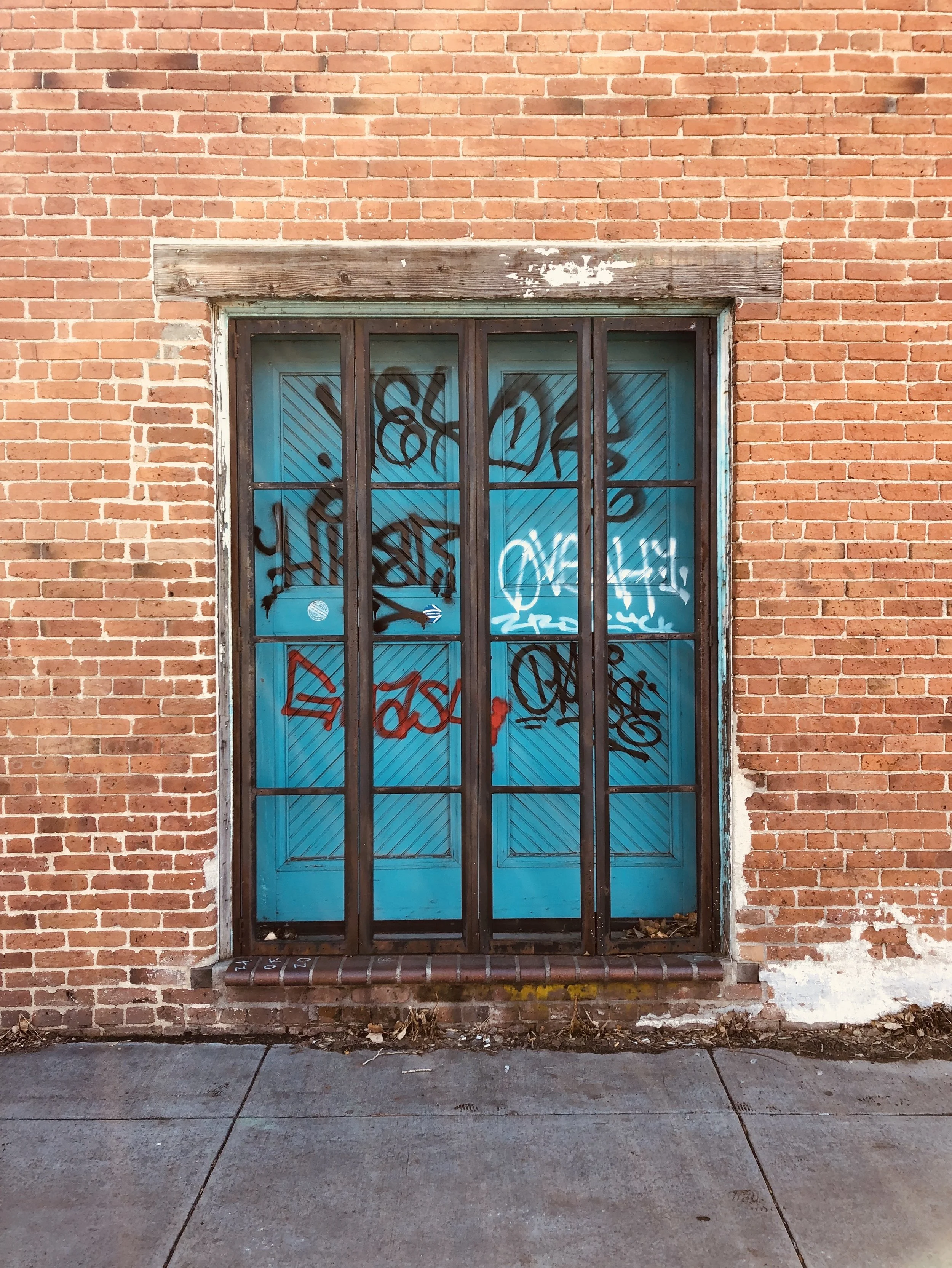 A brick building with a barred blue door covered in graffiti, with a stone step at the bottom and surrounded by a sidewalk and some dirt and leaves.