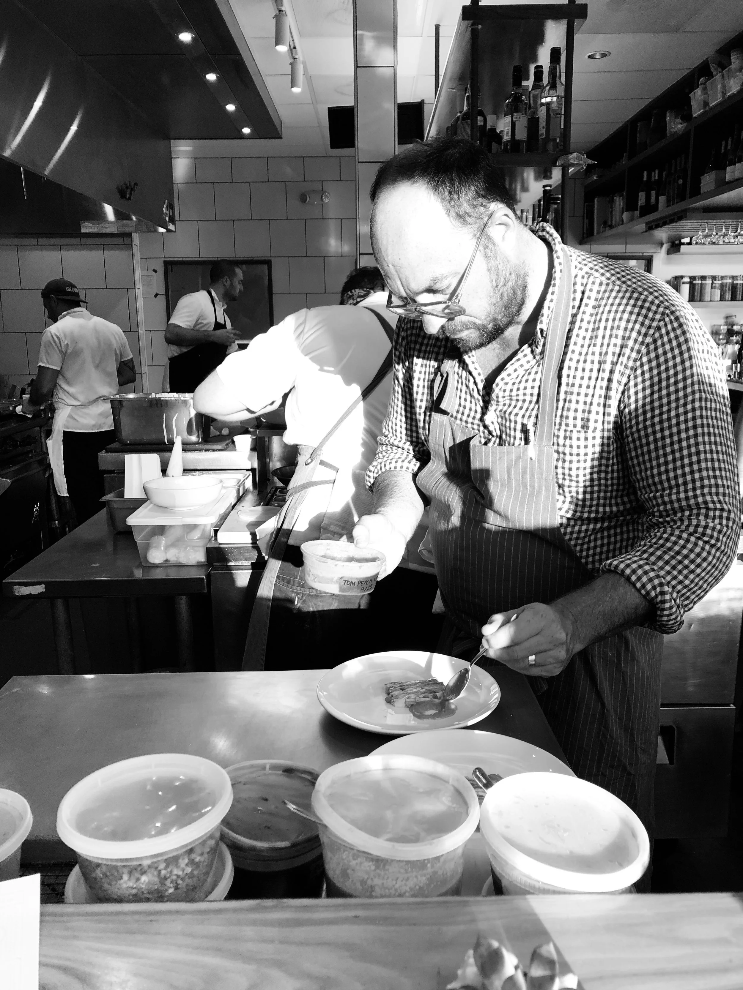 A man with glasses and a checkered shirt preparing food in a restaurant kitchen, with chefs in white and black uniforms working in the background and containers of ingredients on the counter in front.