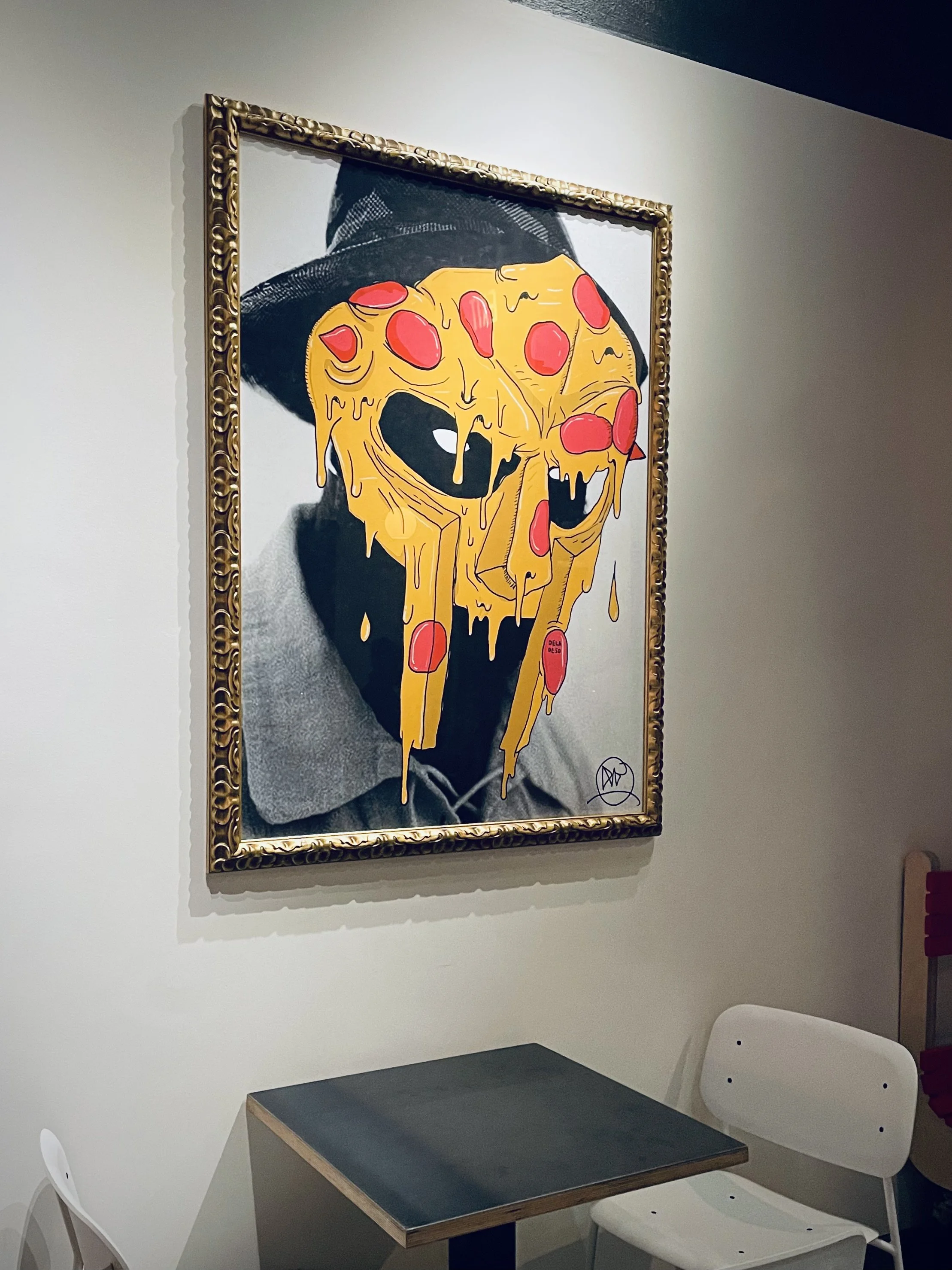 A table with a black top in a room with white walls and two white chairs. A framed artwork hangs on the wall depicting a person wearing a black hat and glasses with a melting pizza slice covering their face.