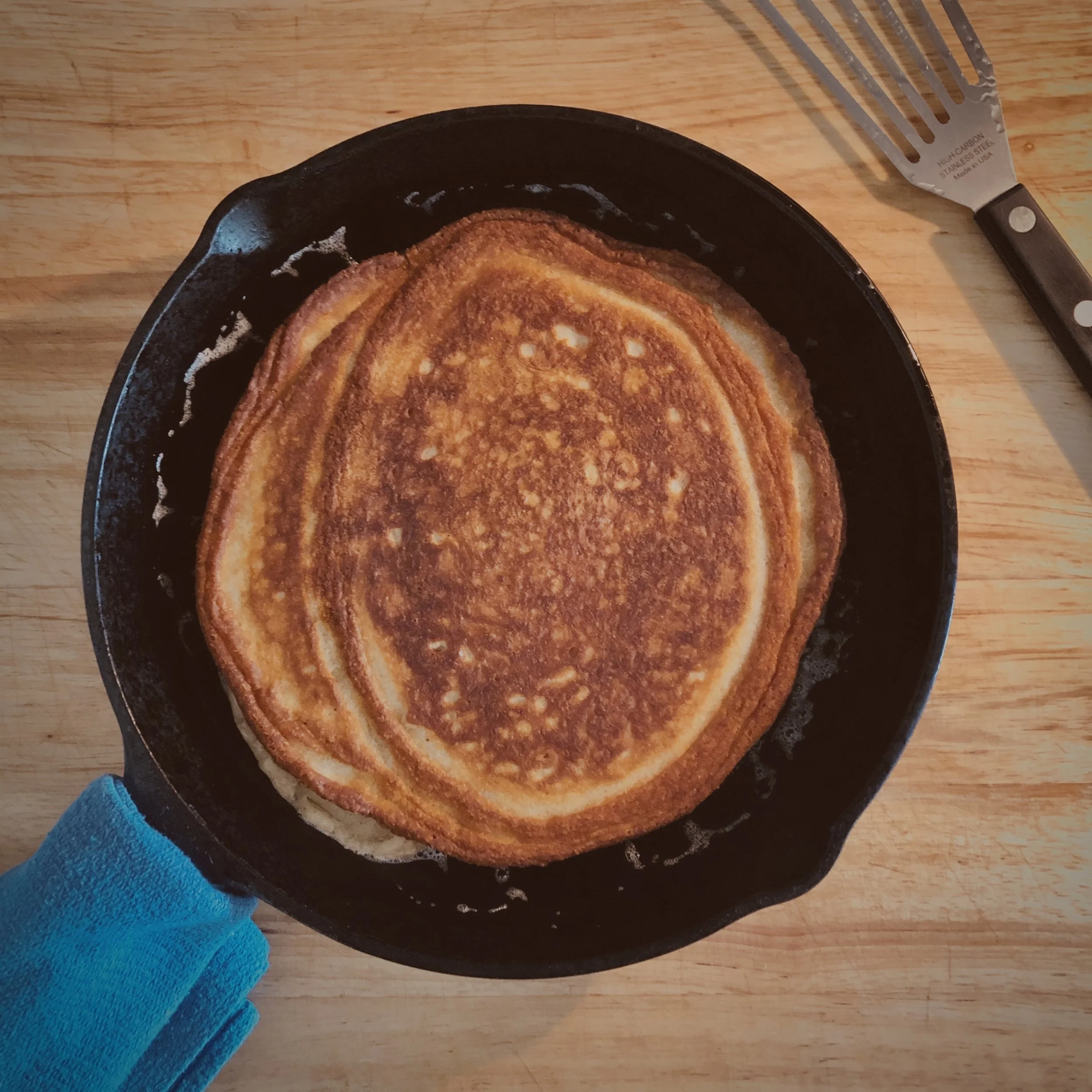 A crispy, golden-brown pancake or crepe in a black skillet on a wooden surface, with a person wearing a blue sleeve holding it.