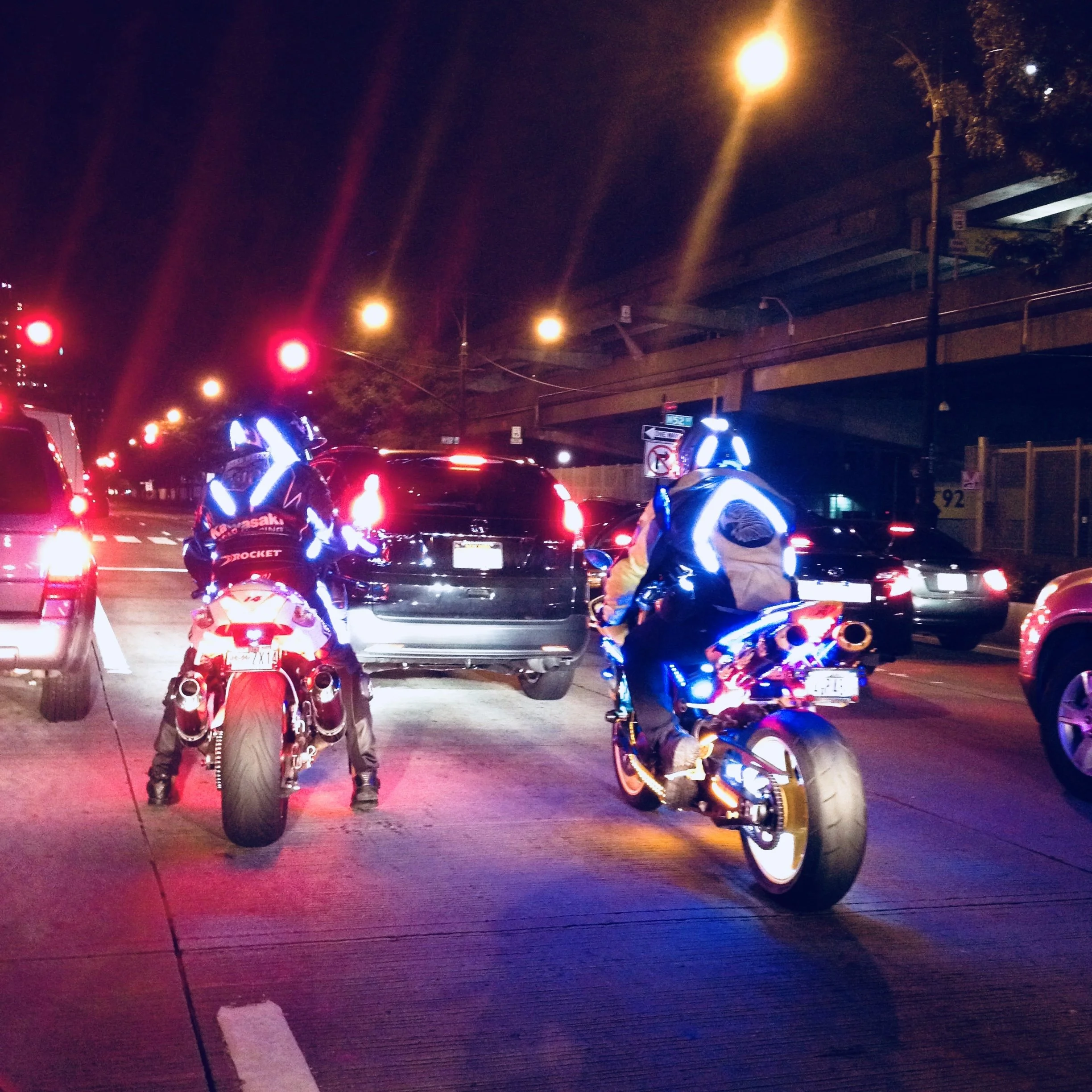 Nighttime scene at a traffic stop with two police officers on motorcycles and several cars waiting at red lights. Streetlights and a bridge are visible in the background.