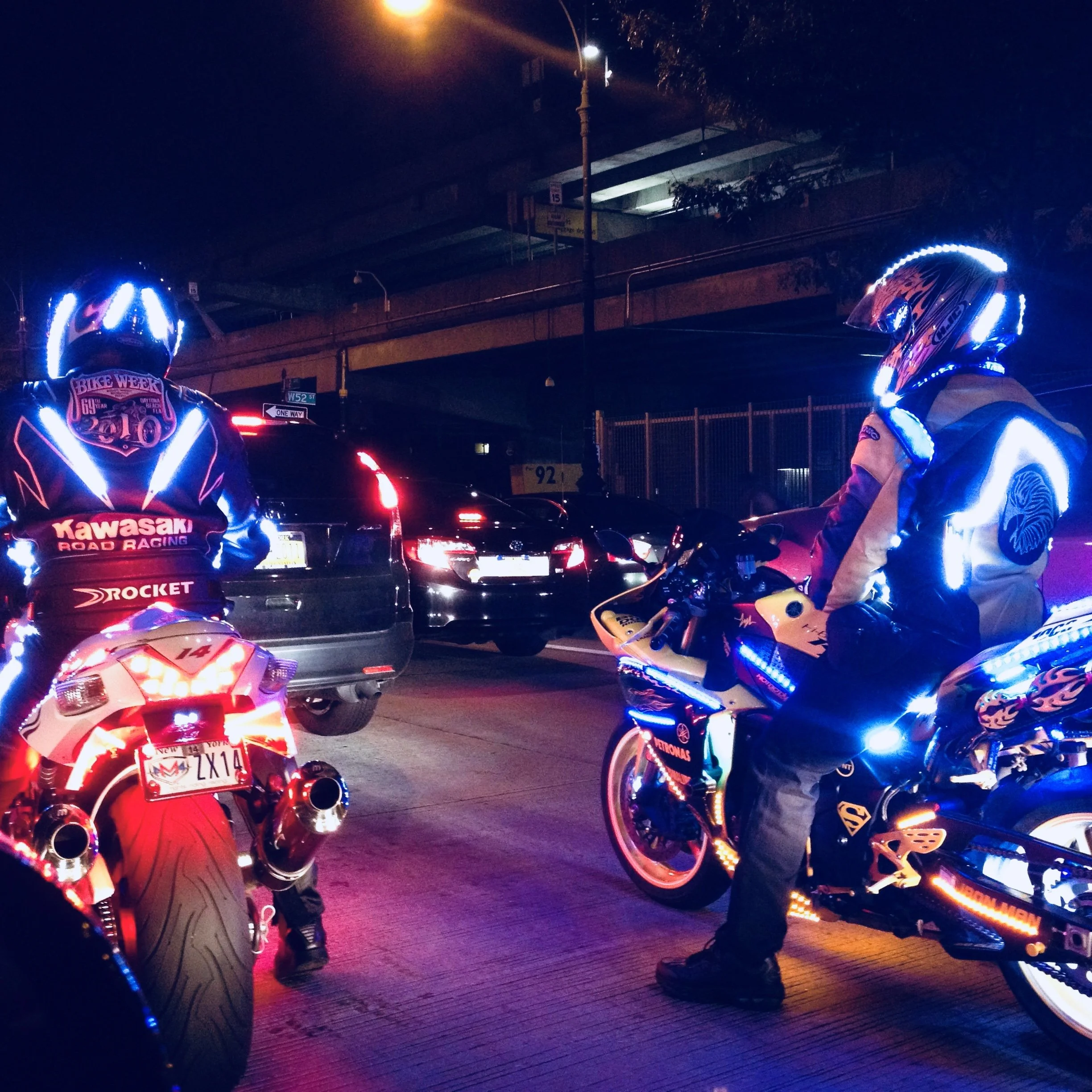Several motorcyclists wearing illuminated helmets and riding gear, gathered at night under a bridge with cars and traffic lights visible in the background.