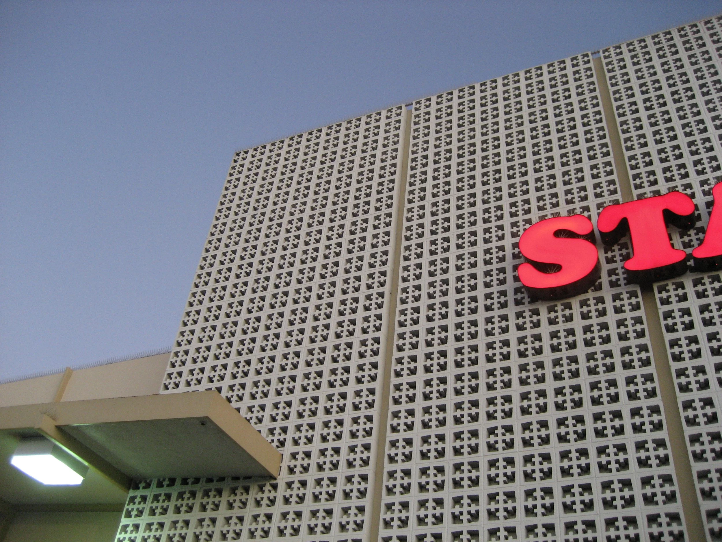 Part of a building with a textured white lattice wall and a red neon sign spelling 'ST'. There is an overhang with a green sign below it and a visible pale blue sky.