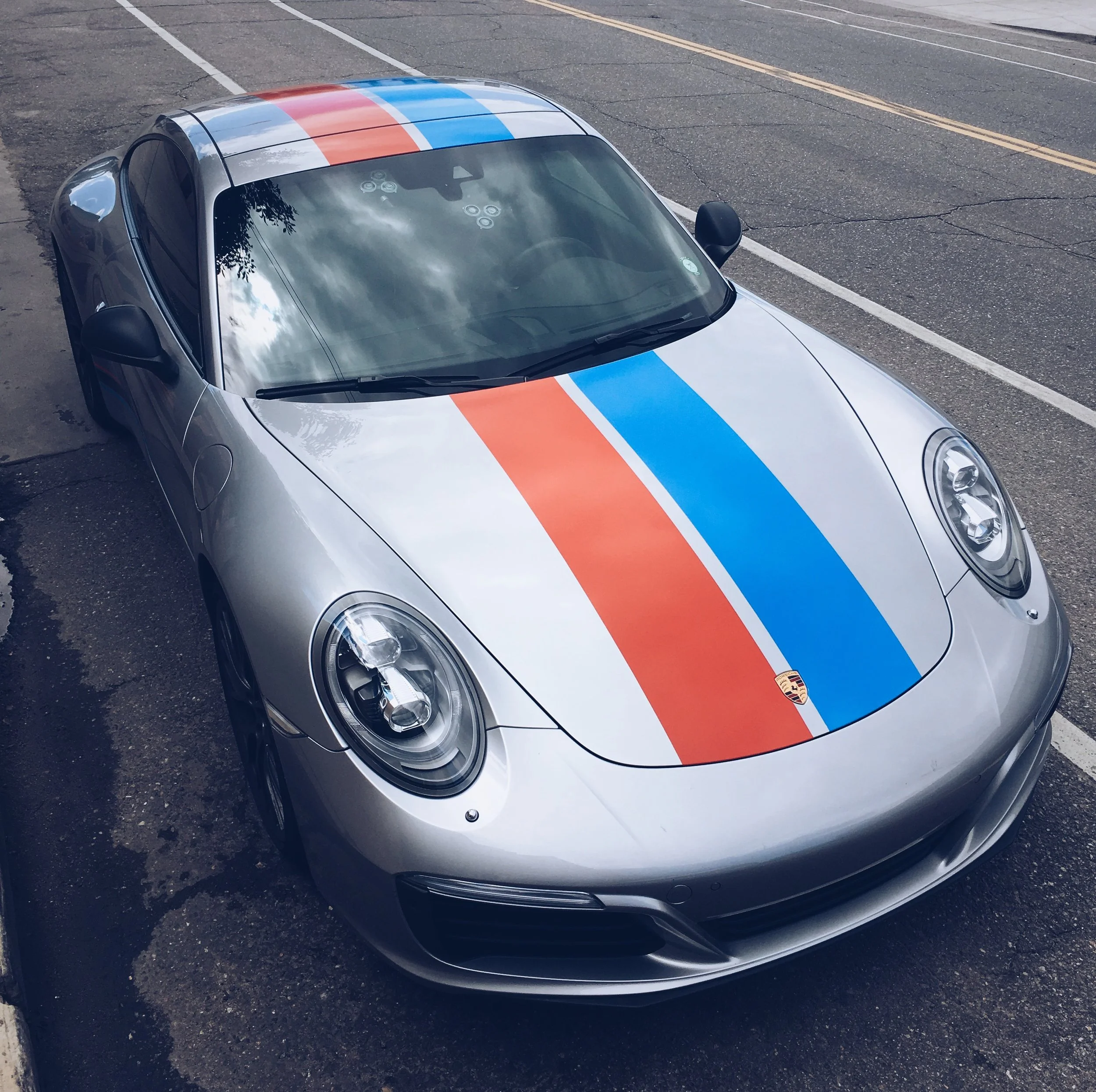 A silver sports car, likely a Porsche, parked in a parking lot. The car has a distinctive red and blue stripe on the hood and roof, and a Porsche emblem on the front.
