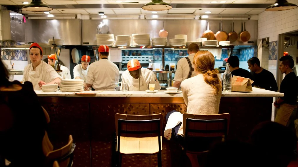 A busy restaurant kitchen with chefs preparing food, a customer sitting at the counter, and other staff working in the background.