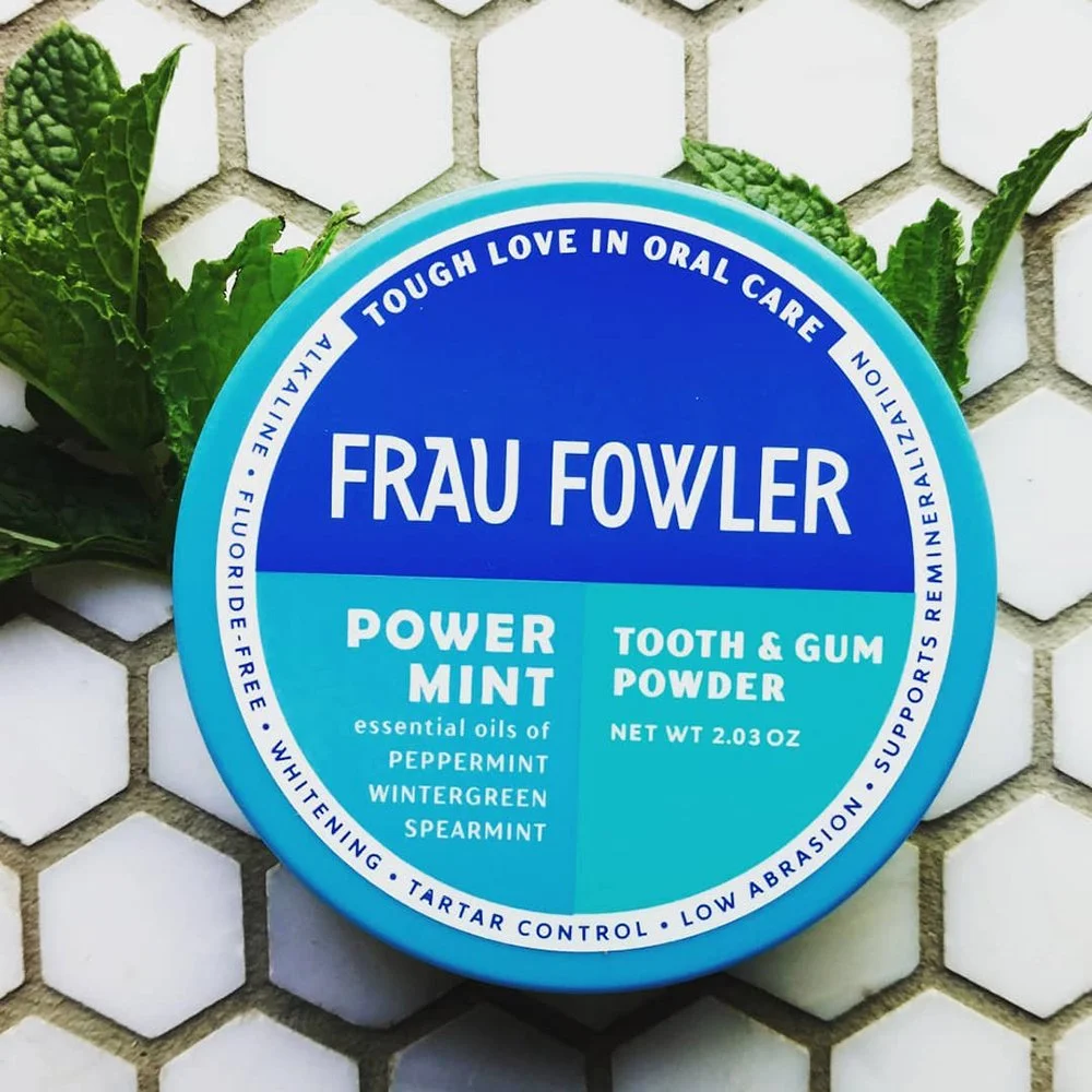 A round container of Frau Fowler Power Mint Tooth & Gum Powder with essential oils of peppermint, wintergreen, and spearmint, placed on a hexagonal white tiled surface with green mint leaves around it.