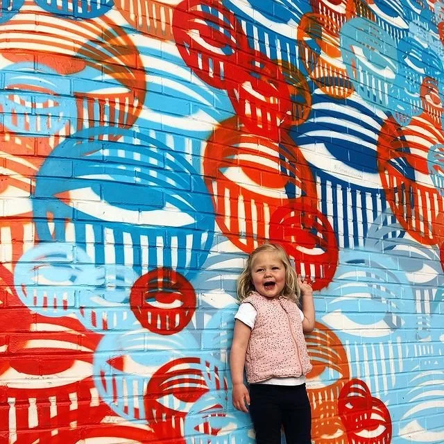 A young girl standing in front of a colorful mural of red, blue, and orange circular patterns with smiling lips and horizontal stripes.