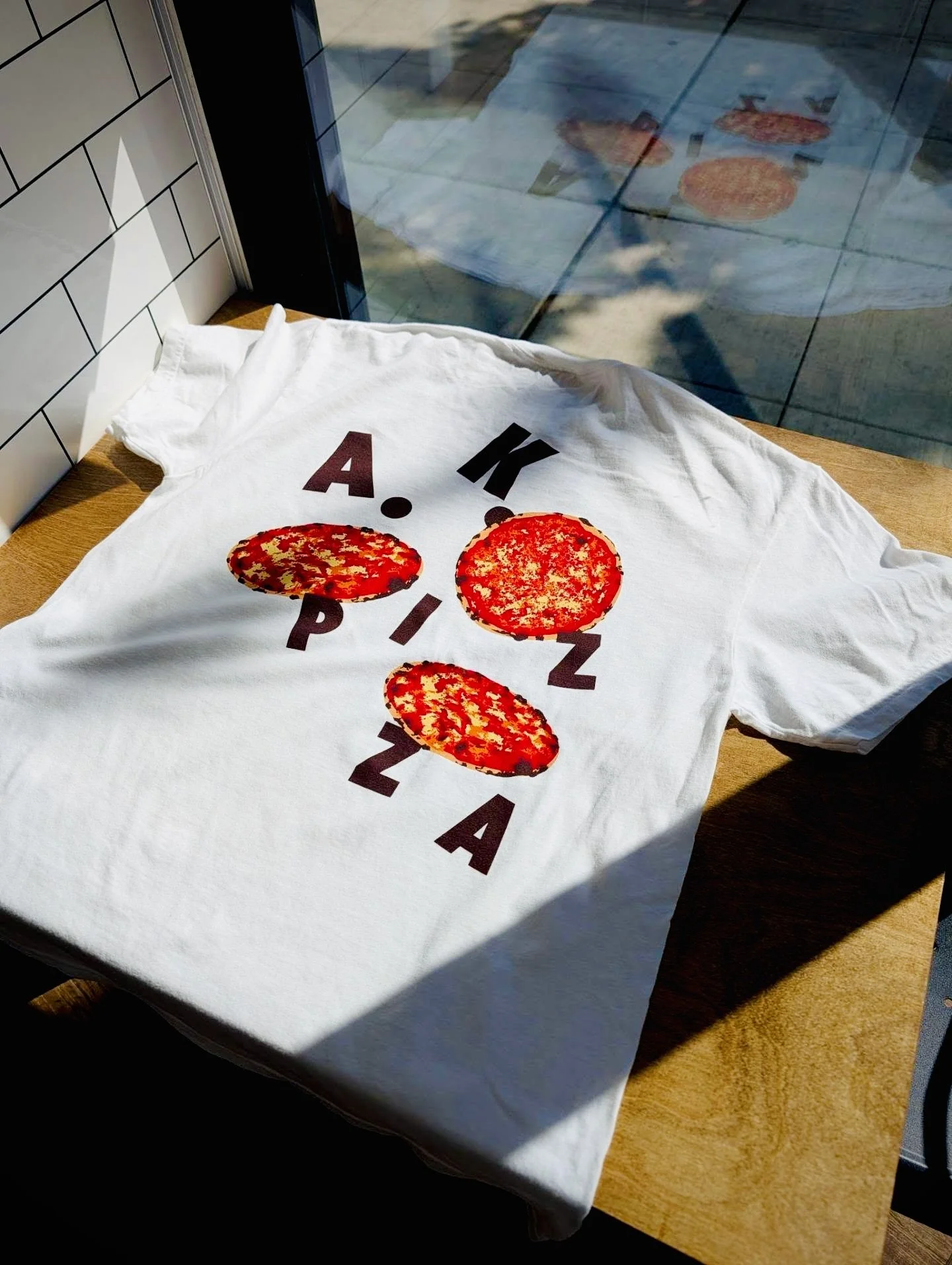A white T-shirt with a design featuring slices of pepperoni pizza arranged vertically, and the text 'A K P I Z A' printed around the slices.