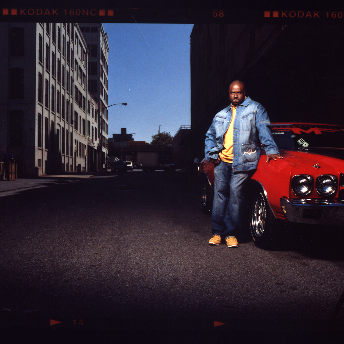 A man standing beside a red classic car on an urban street during daytime, with tall buildings and a clear blue sky in the background.