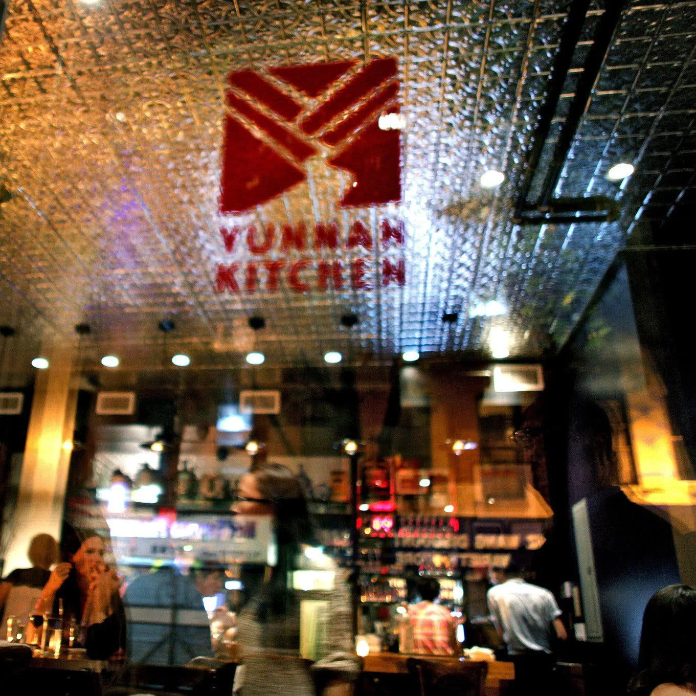 Interior of Vuhann Kitchen restaurant with a glowing sign on the ceiling, patrons dining and socializing, warm lighting, and a reflective ceiling surface.