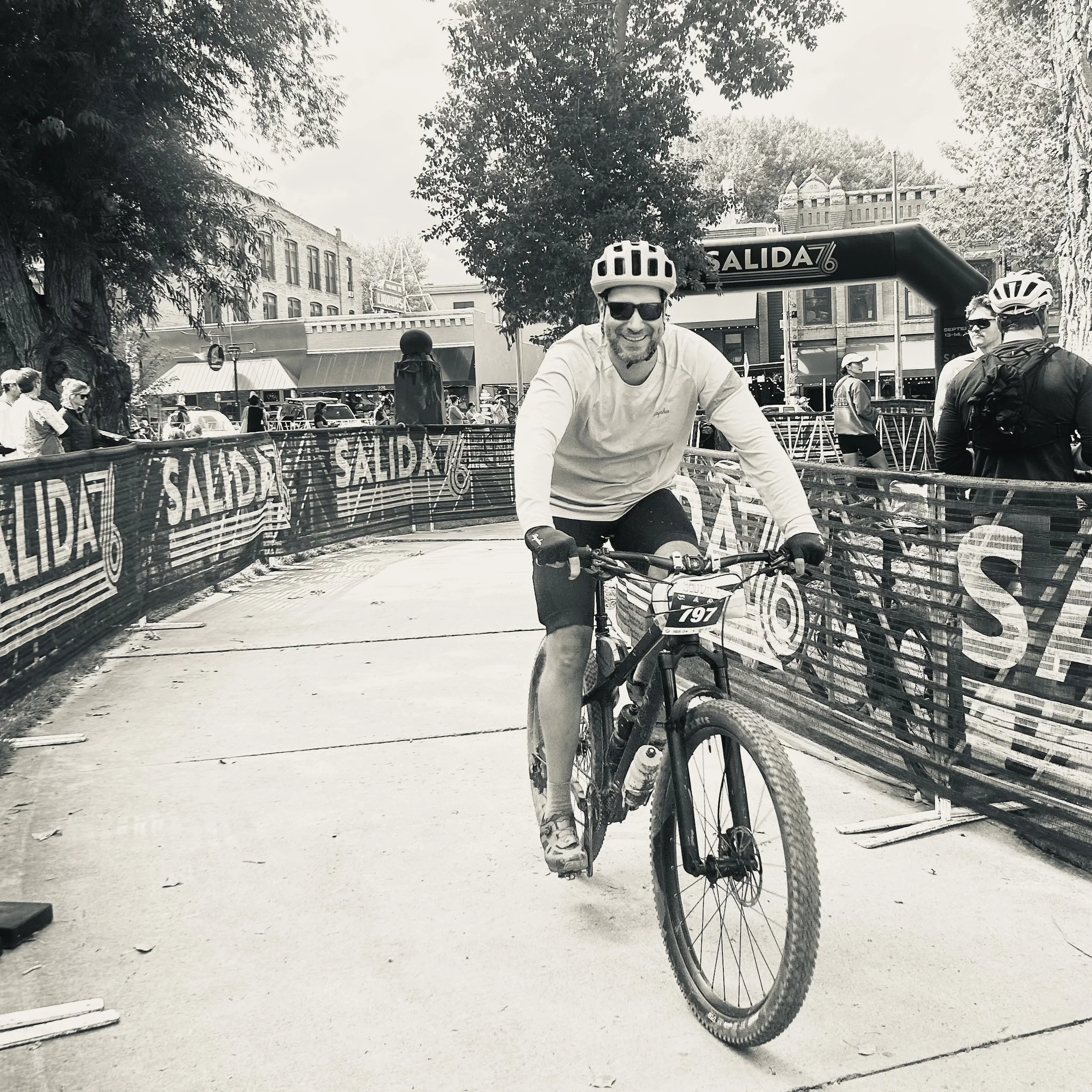 A man riding a bicycle with a helmet and sunglasses on a city street, surrounded by people and trees, with a sign reading 'Salida' overhead.