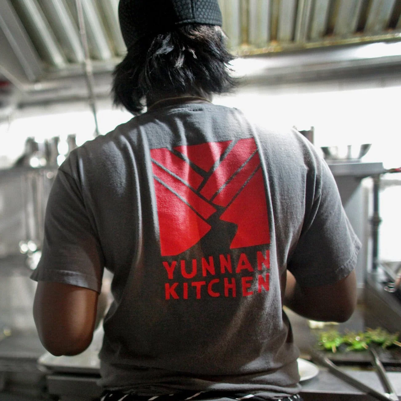Back view of a person wearing a gray T-shirt with a red graphic and text that reads 'Yunnan Kitchen,' working in a kitchen with stainless steel equipment.