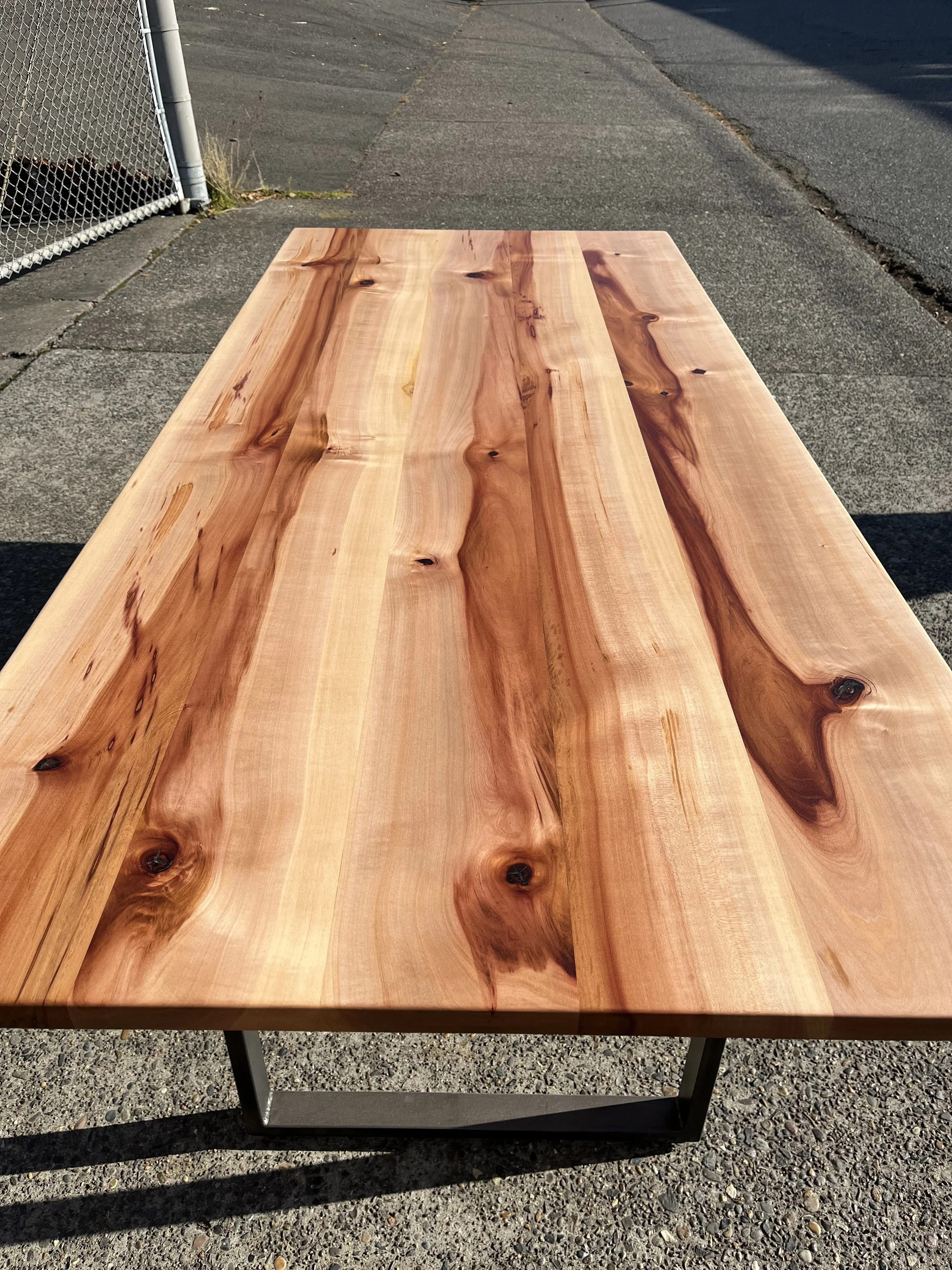 Wooden table with a natural finish outdoors on a sidewalk, next to a chain-link fence.