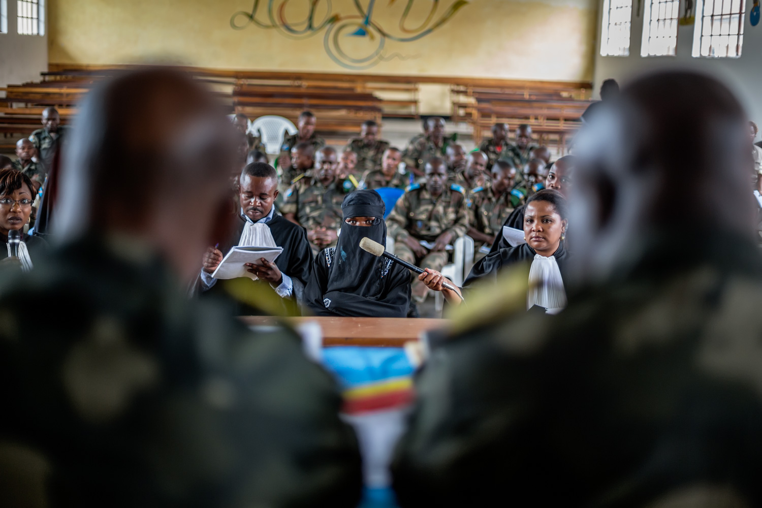  A victim testifies before the closed military tribunal. A member of the prosecution team holds a microphone up for her while a defense lawyer makes notes. The accused soldiers are seated in the rear. Victims are often extremely reluctant to step for