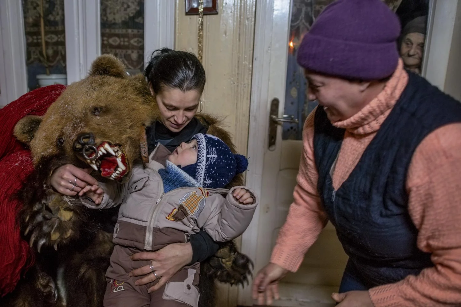 A lady bear in Toloaca's troupe plays with the young son of Cătălin Apetroaie, a bear dancer himself, after a performance at Apetroaie's home. The child's grandmother scurries by while his great-grandmother peers from inside the house. December 28, 2014. Laloaia village, Bacău county, Romania.