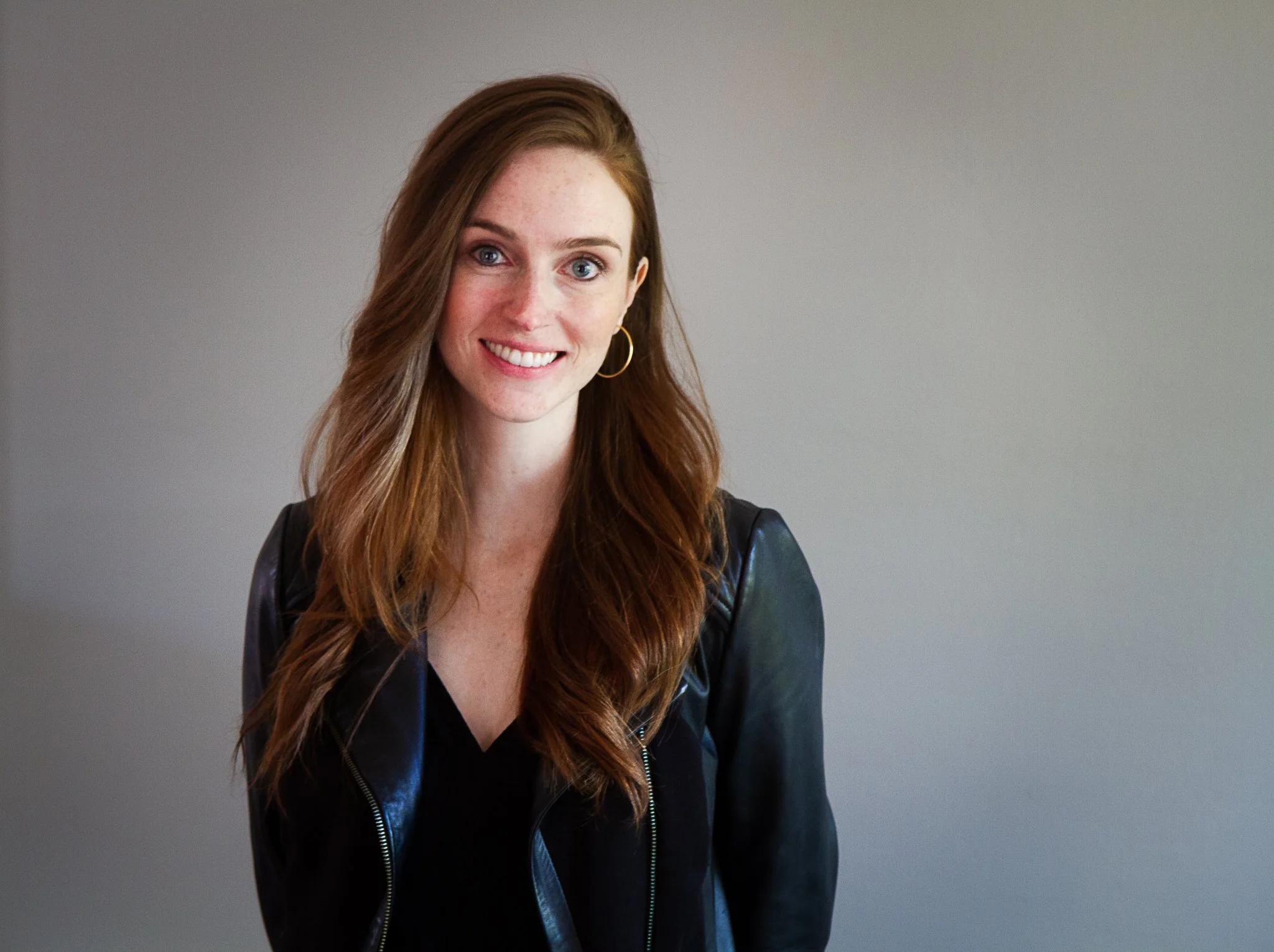 A young woman with long red hair, blue eyes, wearing a black outfit and a black leather jacket, smiling against a neutral background.