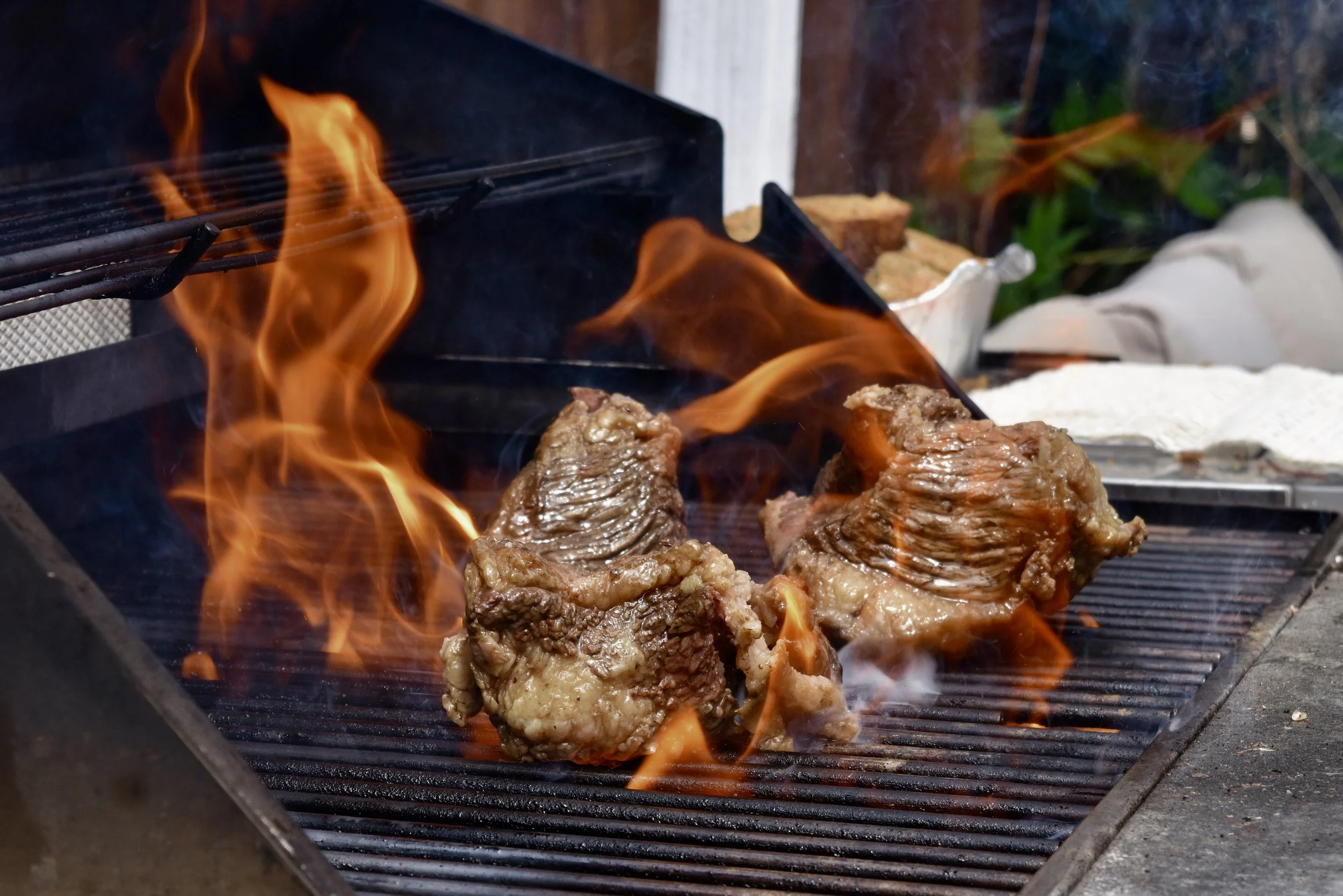 Three pieces of beef being grilled on an outdoor barbecue with flames around.