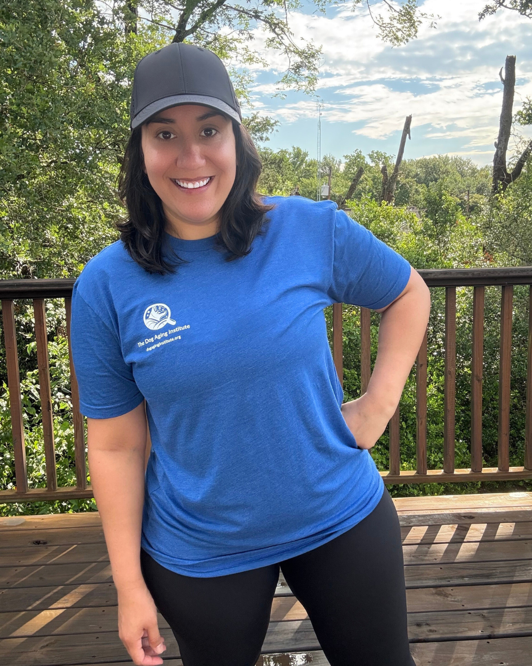 A person wearing a blue t-shirt and a black baseball cap stands on a wooden deck with trees and a cloudy sky in the background.