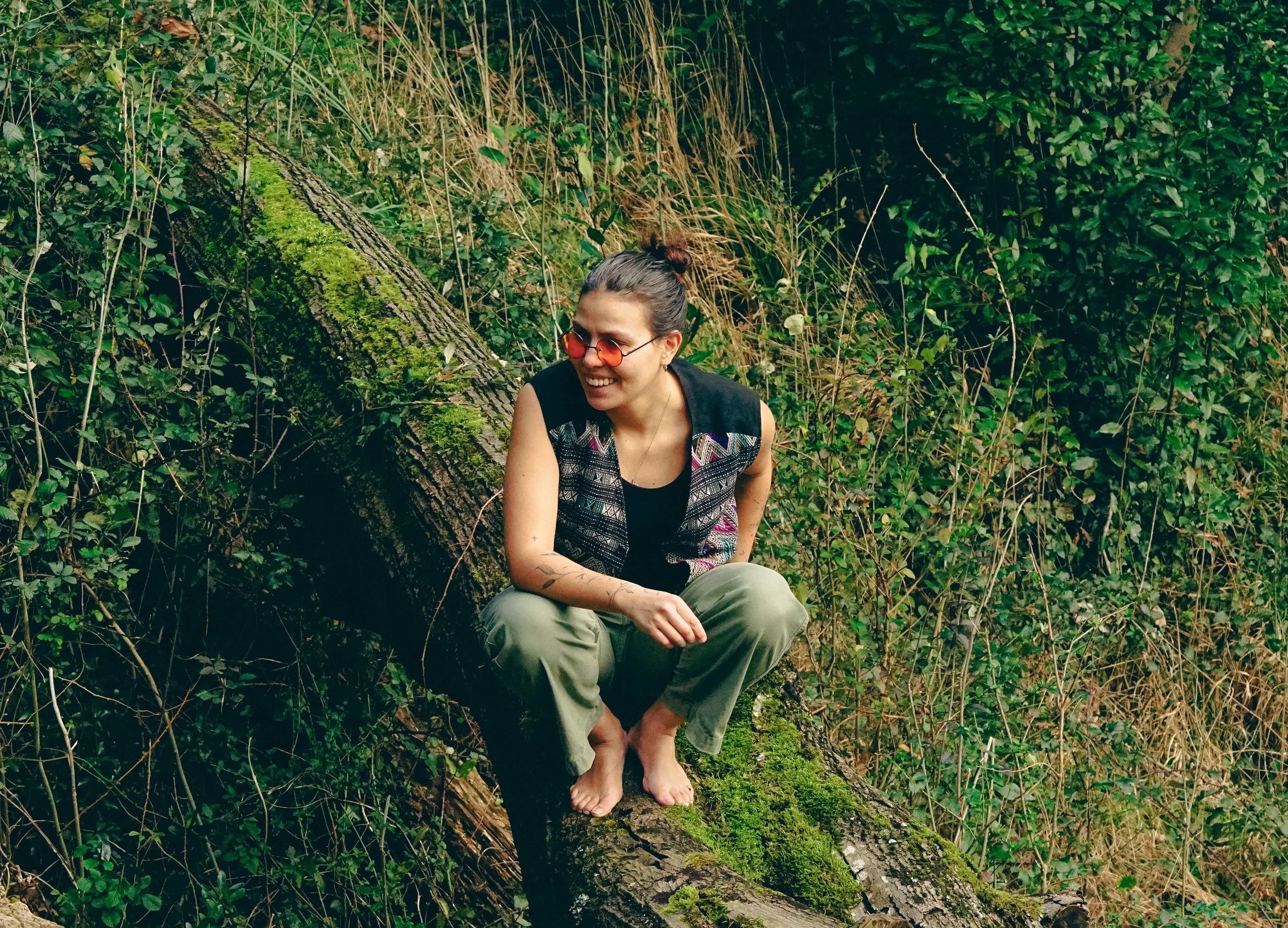 A woman sitting barefoot on a moss-covered fallen tree trunk in a lush green forest, smiling and wearing red-tinted sunglasses and casual outdoor clothing.
