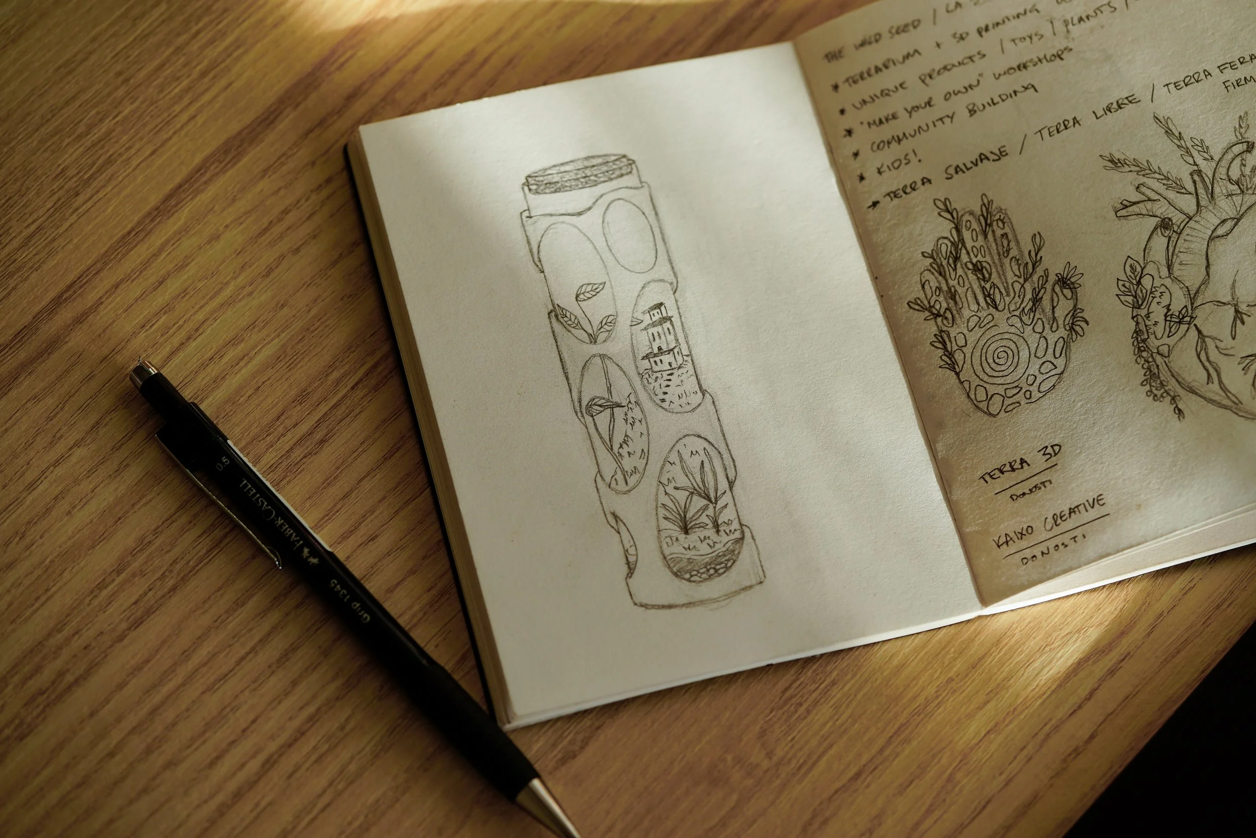 Open sketchbook with a pen on a wooden surface. The left page has a drawing of a tall container with various landscape illustrations inside it. The right page has handwritten notes and additional sketches of plants and a heart-shaped design.