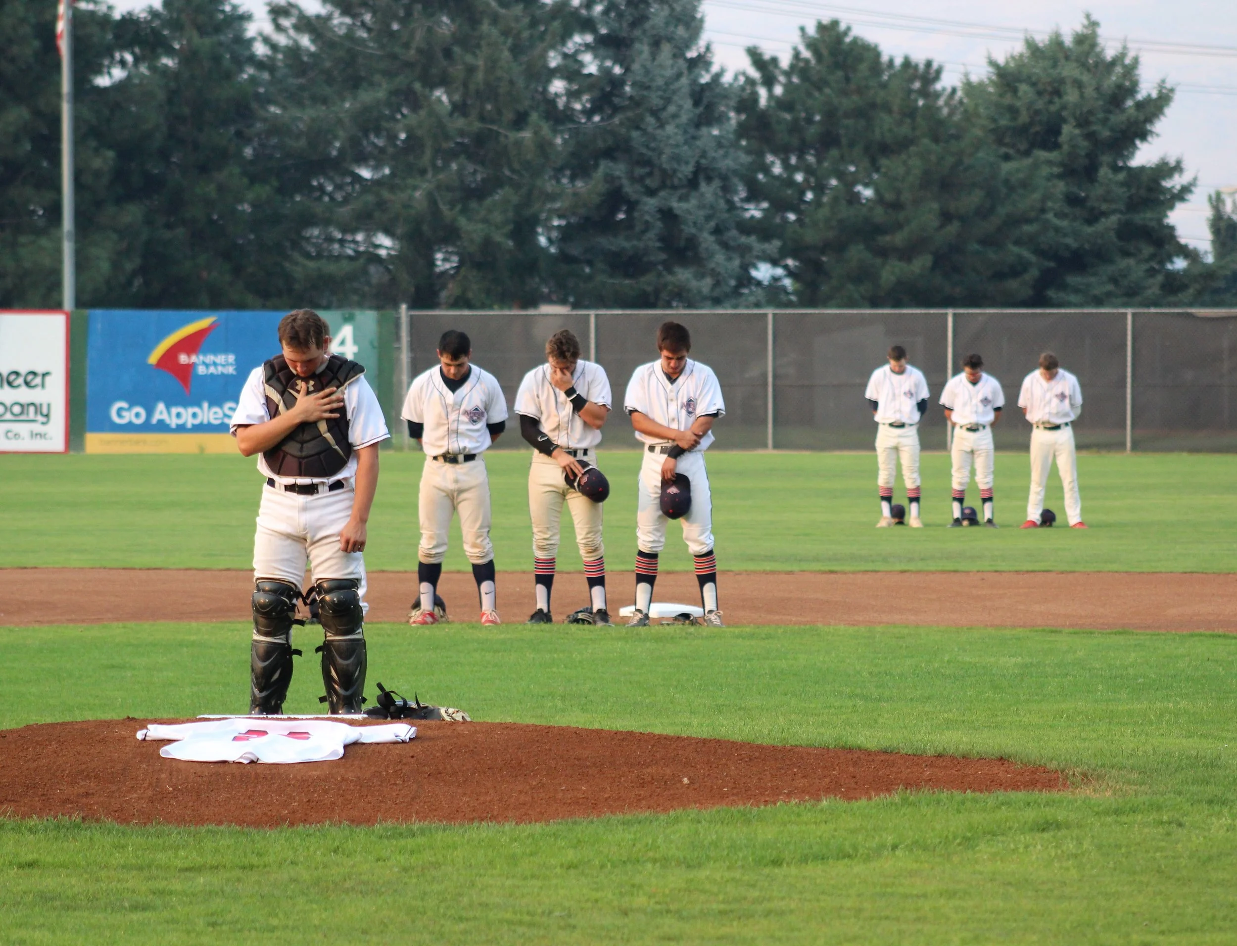 AppleSox players take pause for a moment of silence, Monday night. Wenatchee honored teammate Tommy Watanabe who passed away unexpectedly, Saturday, after playing with the AppleSox through July 22. Watanabe was the team's starting pitcher in the final game of 2017, and they brought his jersey to the mound for the moment of silence and National Anthem. Photo by Alyssa Taylor.