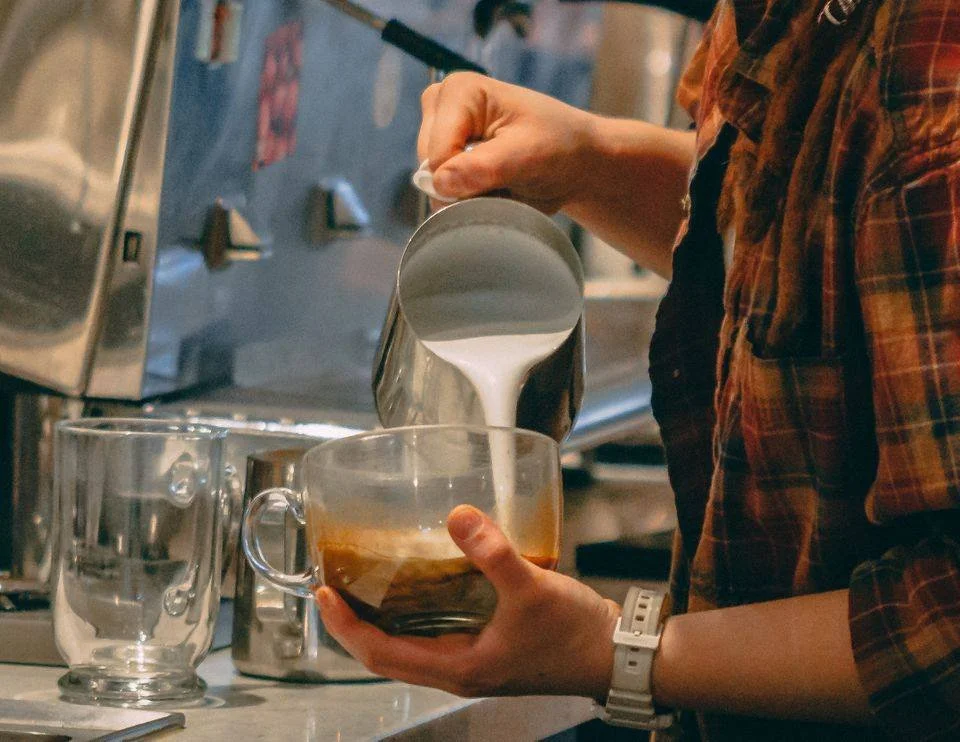  Barista pouring steamed milk into a glass mug of espresso at BeWell Bakery &amp; Cafe. 