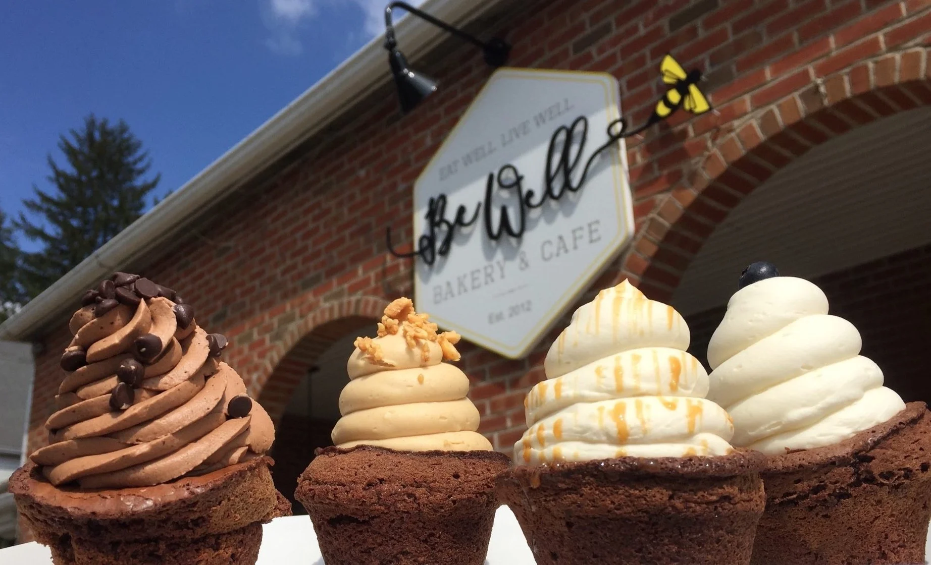  Freshly baked cupcakes with swirled frosting displayed in front of the BeWell Bakery &amp; Cafe storefront sign. 