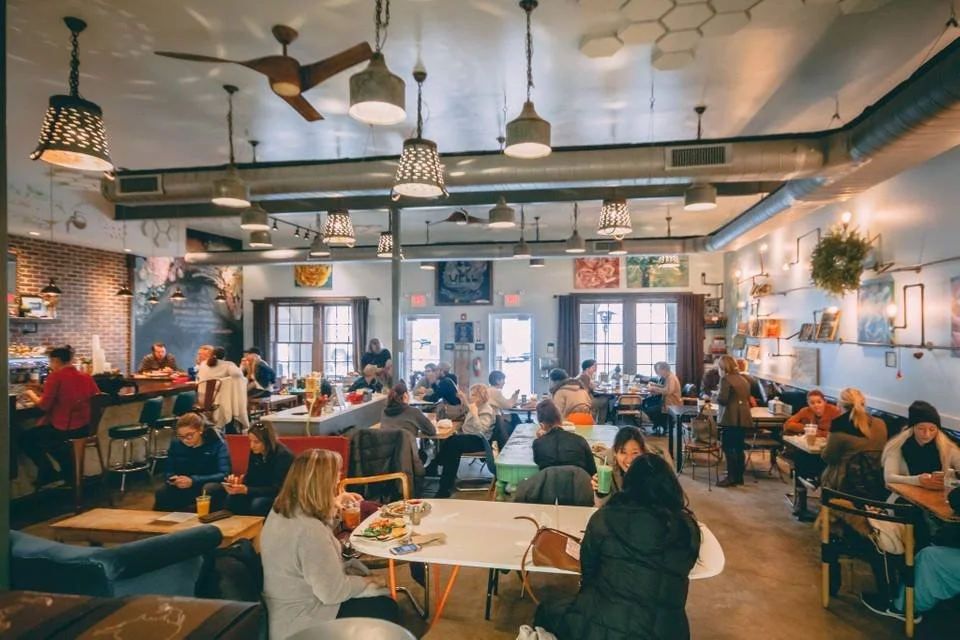  Bright, bustling interior of BeWell Bakery &amp; Cafe with customers enjoying meals around tables. 