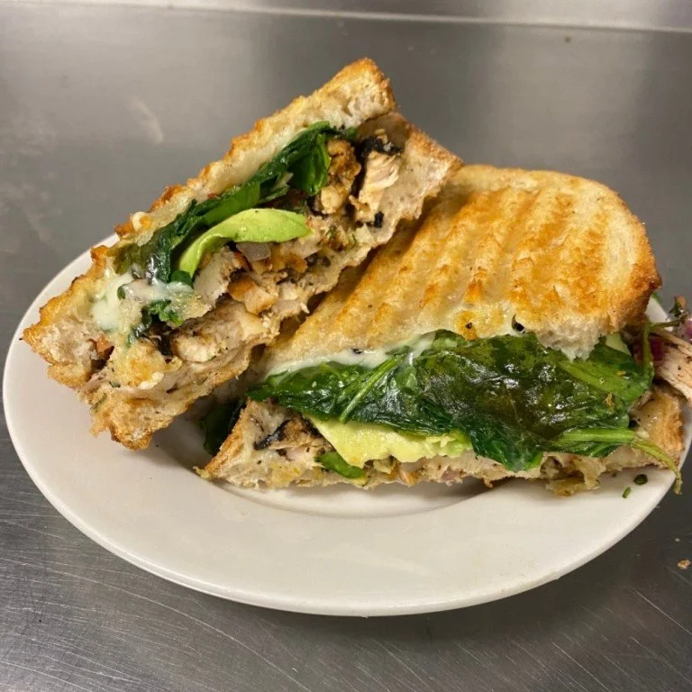  Toasted panini sandwich filled with leafy greens, melted cheese, and vegetables, served at BeWell Bakery &amp; Cafe. 