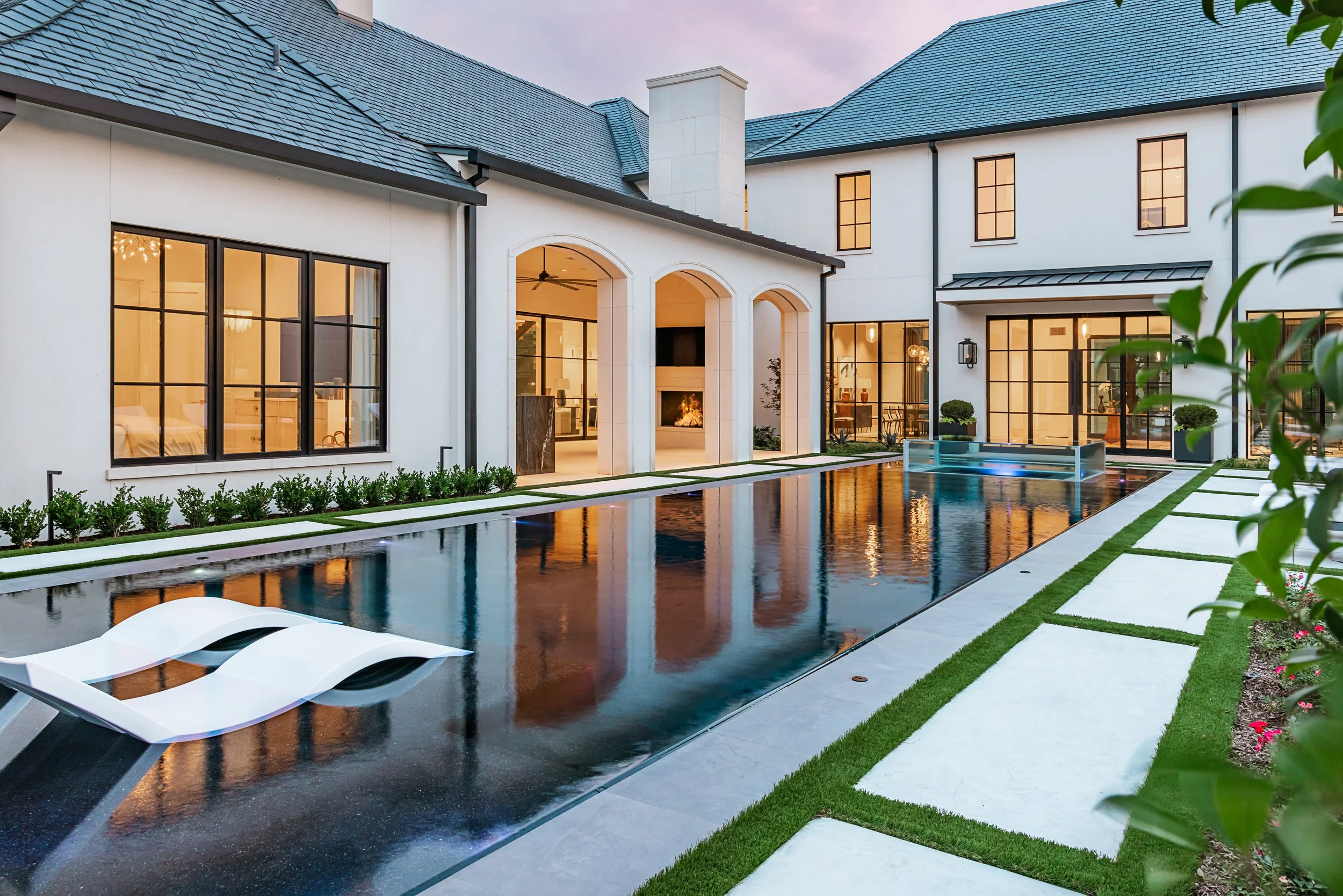 Luxury house with a swimming pool in the backyard, modern lounge chairs, and large glass windows with warm lighting inside, surrounded by well-maintained lawn and greenery.
