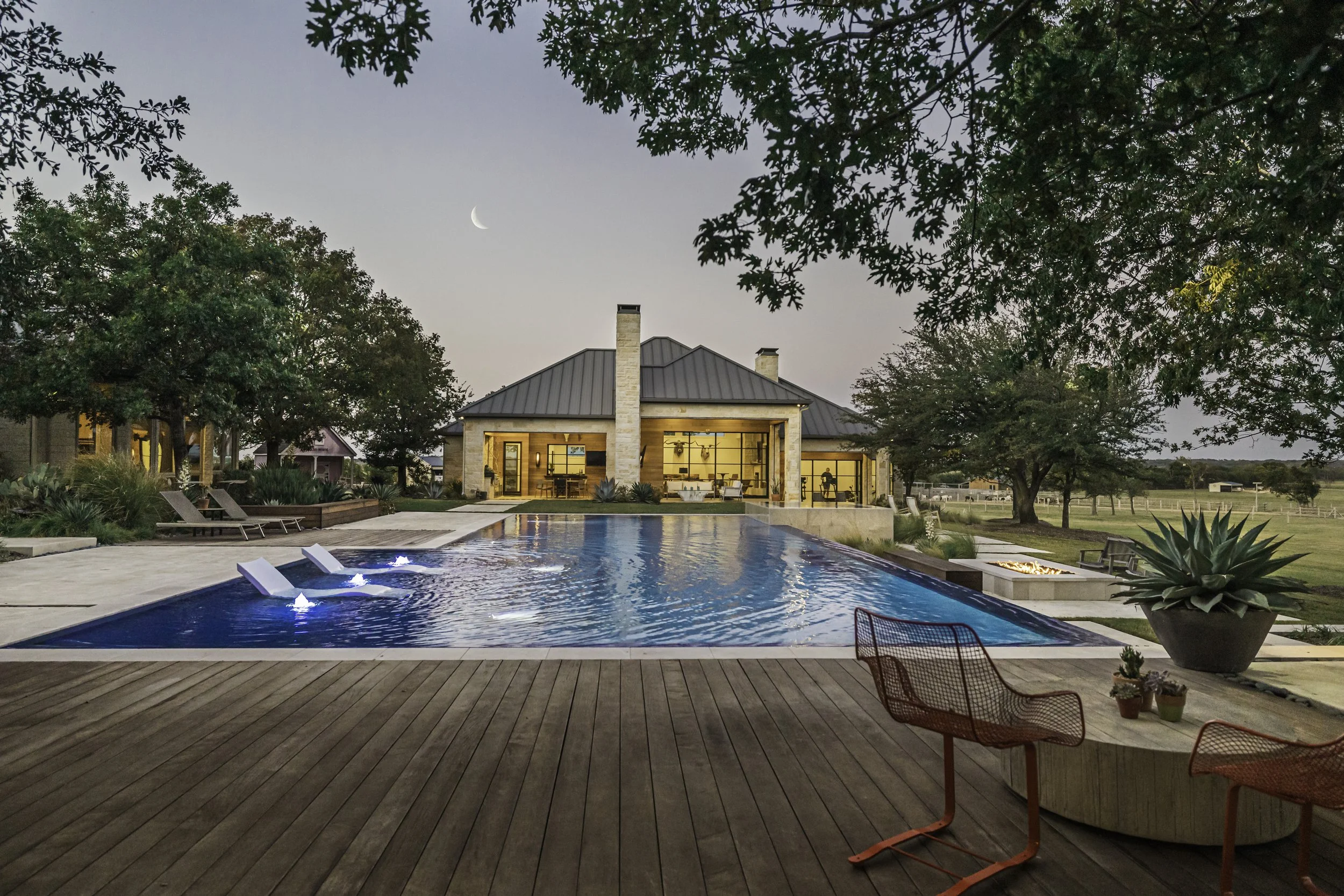 A backyard with a swimming pool, surrounded by a wooden deck, outdoor furniture, green trees, and a house with large glass doors and windows at dusk. The moon is visible in the sky.