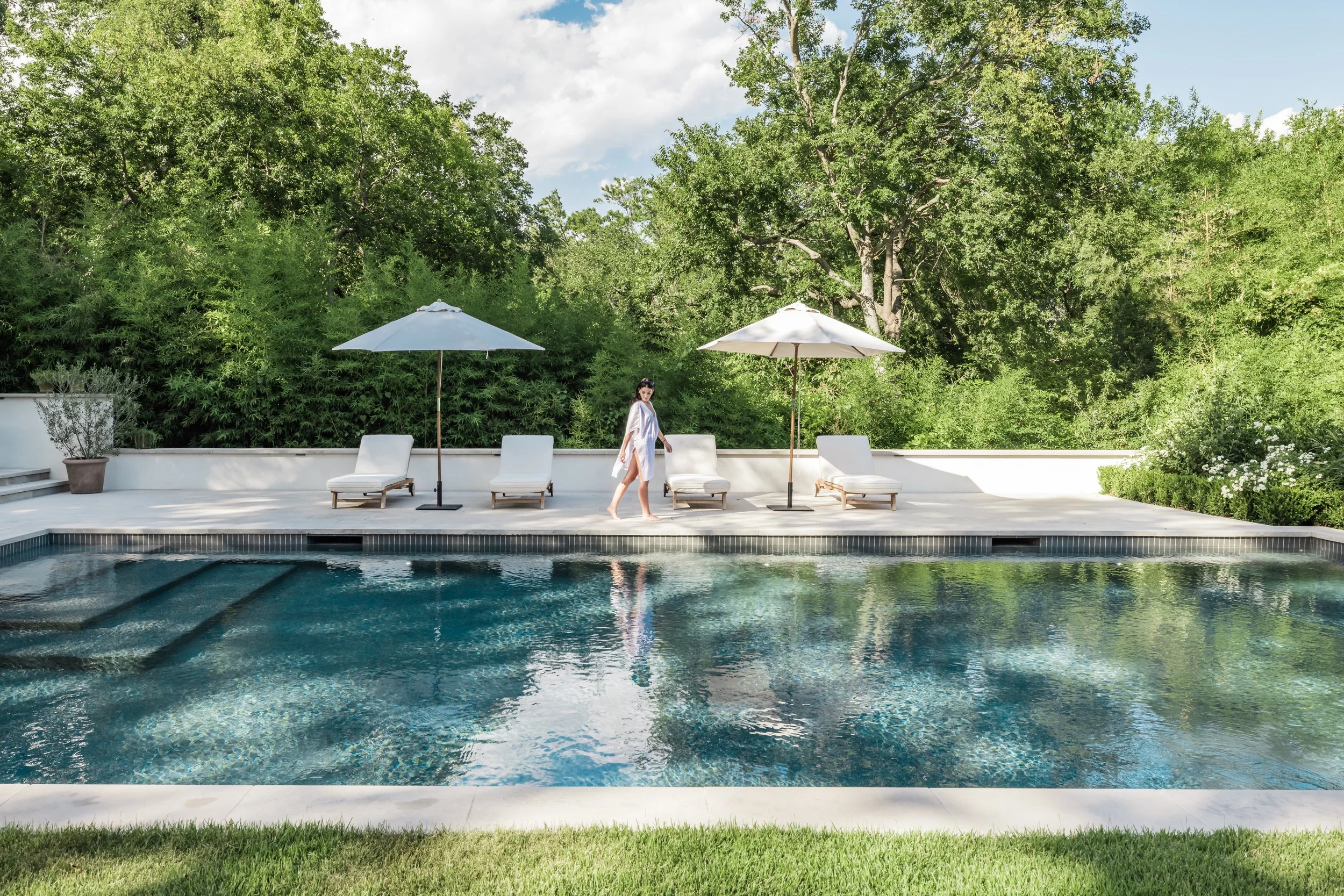 A woman in a white cover-up walking by a poolside with lounge chairs and umbrellas, with a background of lush green trees and a partly cloudy sky.