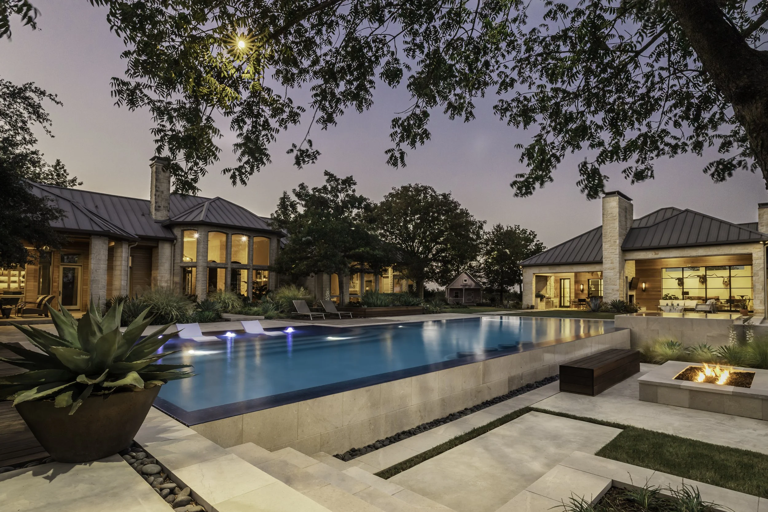 A modern house with a lit interior and large windows, a swimming pool in the backyard with underwater lighting, landscaped garden with plants and trees, outdoor fire pit, and a clear evening sky with the sun setting.