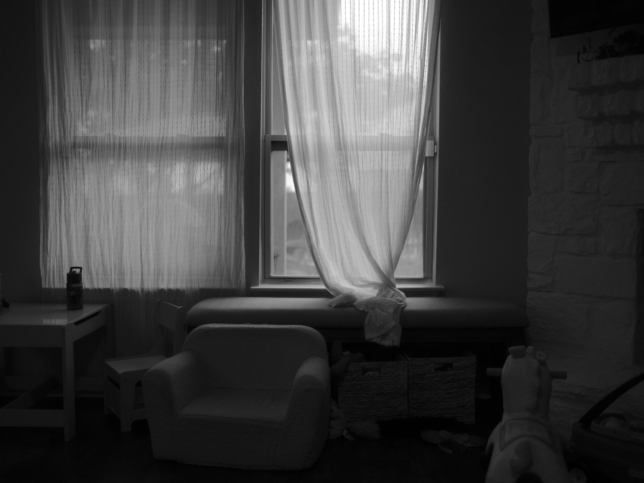 Black and white image of a room with two windows covered by sheer curtains, a small chair, a bench with storage baskets underneath, and a toy horse on wheels.