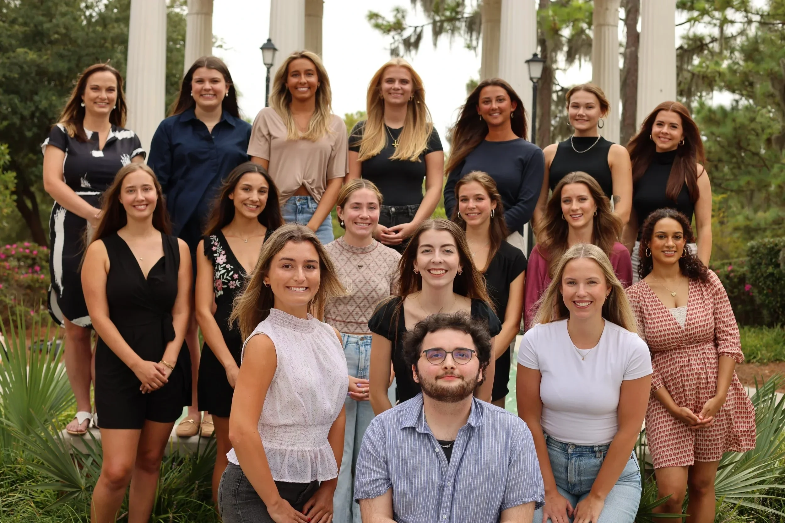 A group of 19 young women and one young man posing outdoors in front of a white pavilion with columns and greenery. The group is arranged in four rows, with the man seated in the front.
